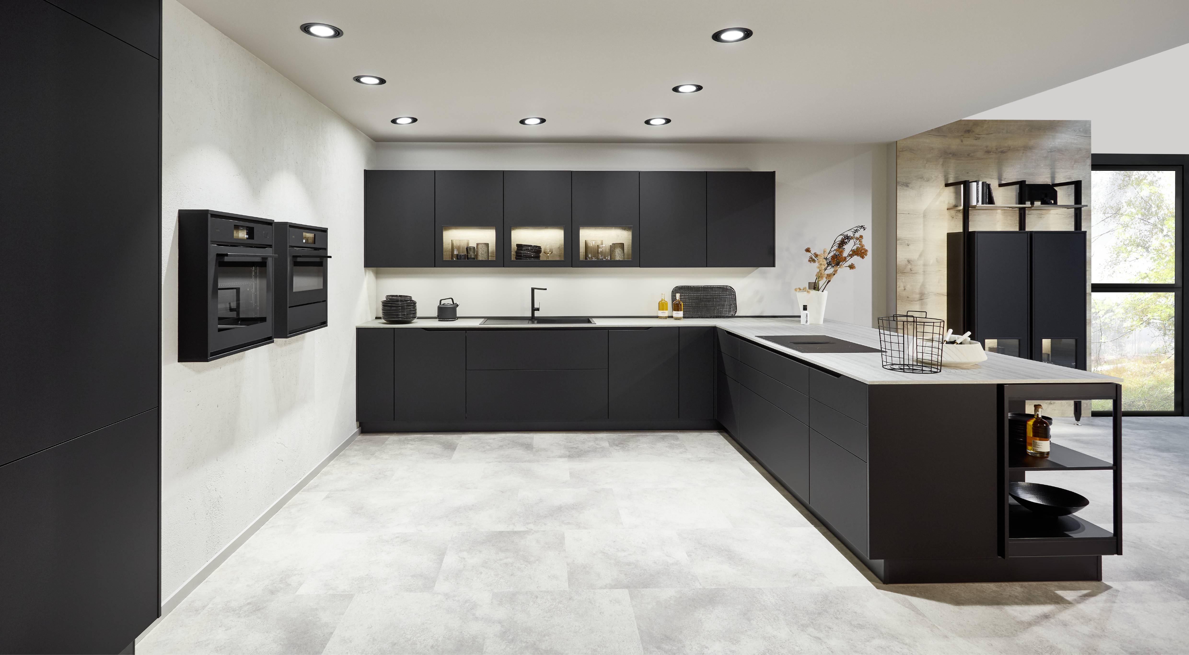 Uncompromising in every detail — the Seno kitchen in full matte black with illuminated glass upper cabinets, a wall-mounted appliance column, and an open-shelf island extension delivers a design that holds nothing back.