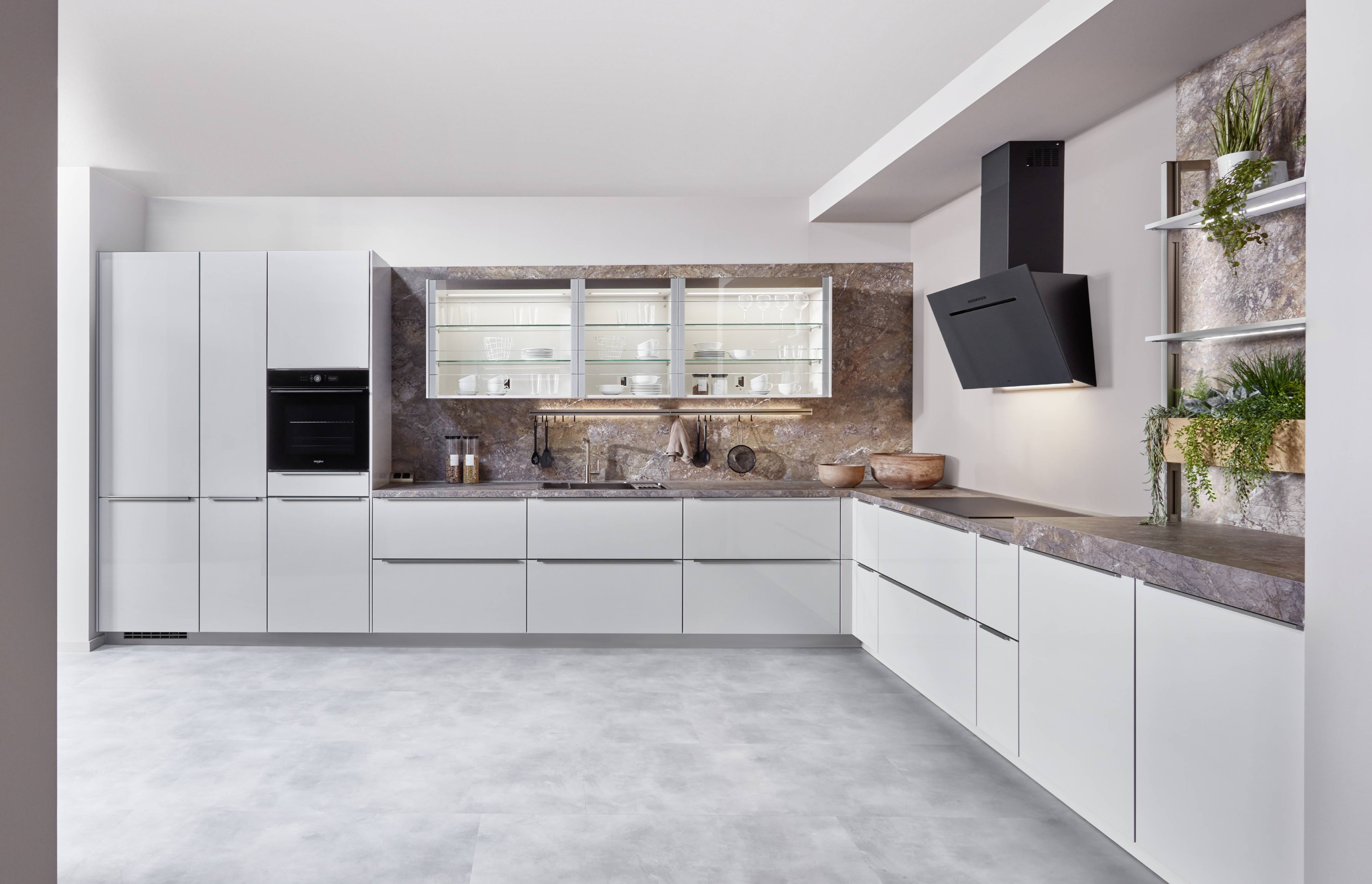 White Cabinetry with Stone Backsplash and Illuminated Glass Wall Cabinet Large L-shaped Novalux kitchen with matte white handleless cabinetry, a dramatic veined brown and grey stone backsplash and matching worktop, illuminated glass wall cabinet with crockery display, black integrated oven, black angled wall-mounted hood, open shelving with indoor plants, and a utensil rail on the stone backsplash.
