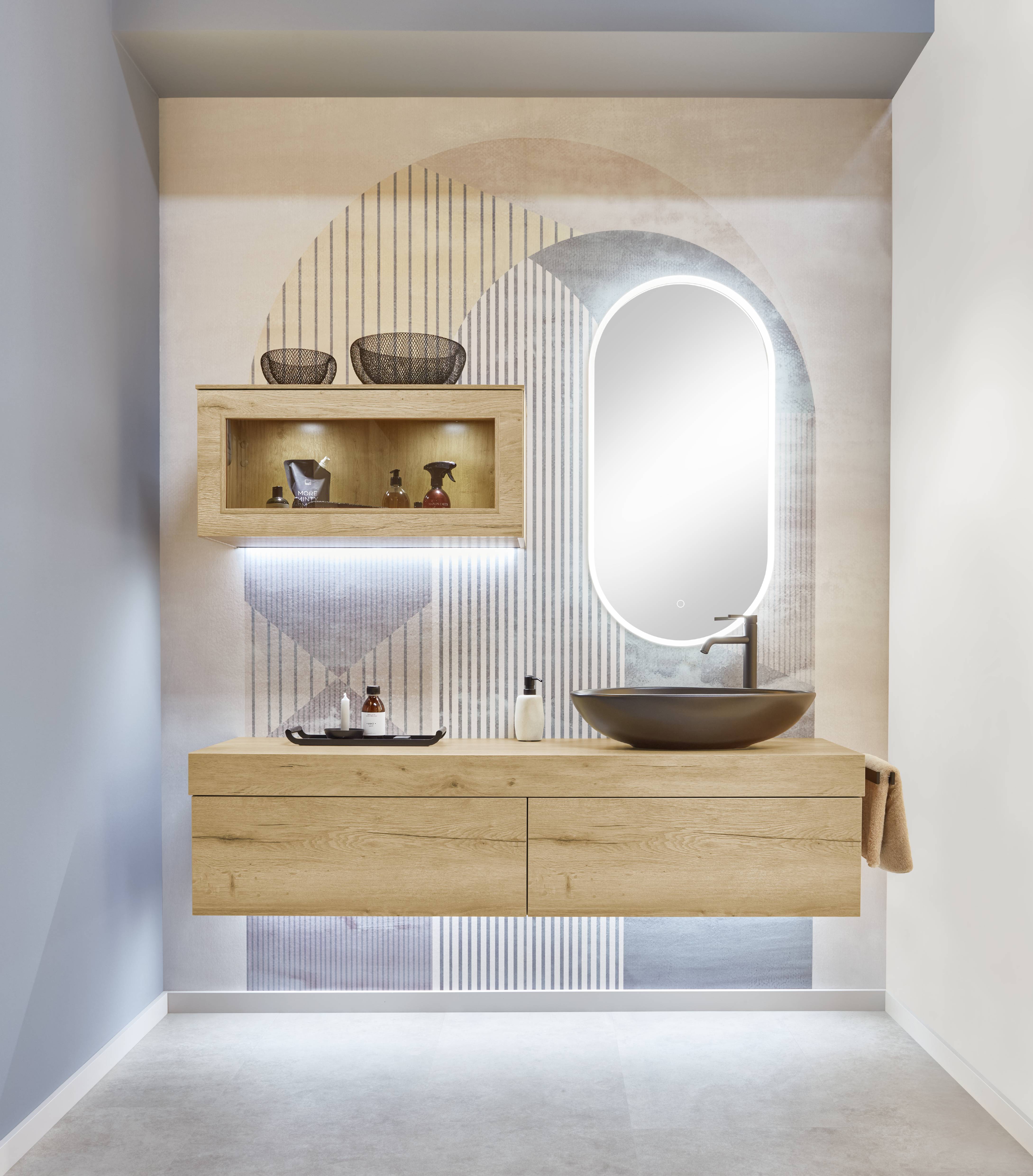 Floating oak bathroom vanity with two drawers, an illuminated oval mirror, an open wall-mounted display cabinet with toiletries, and a matte black vessel sink, set against a patterned arch-motif wallpaper.