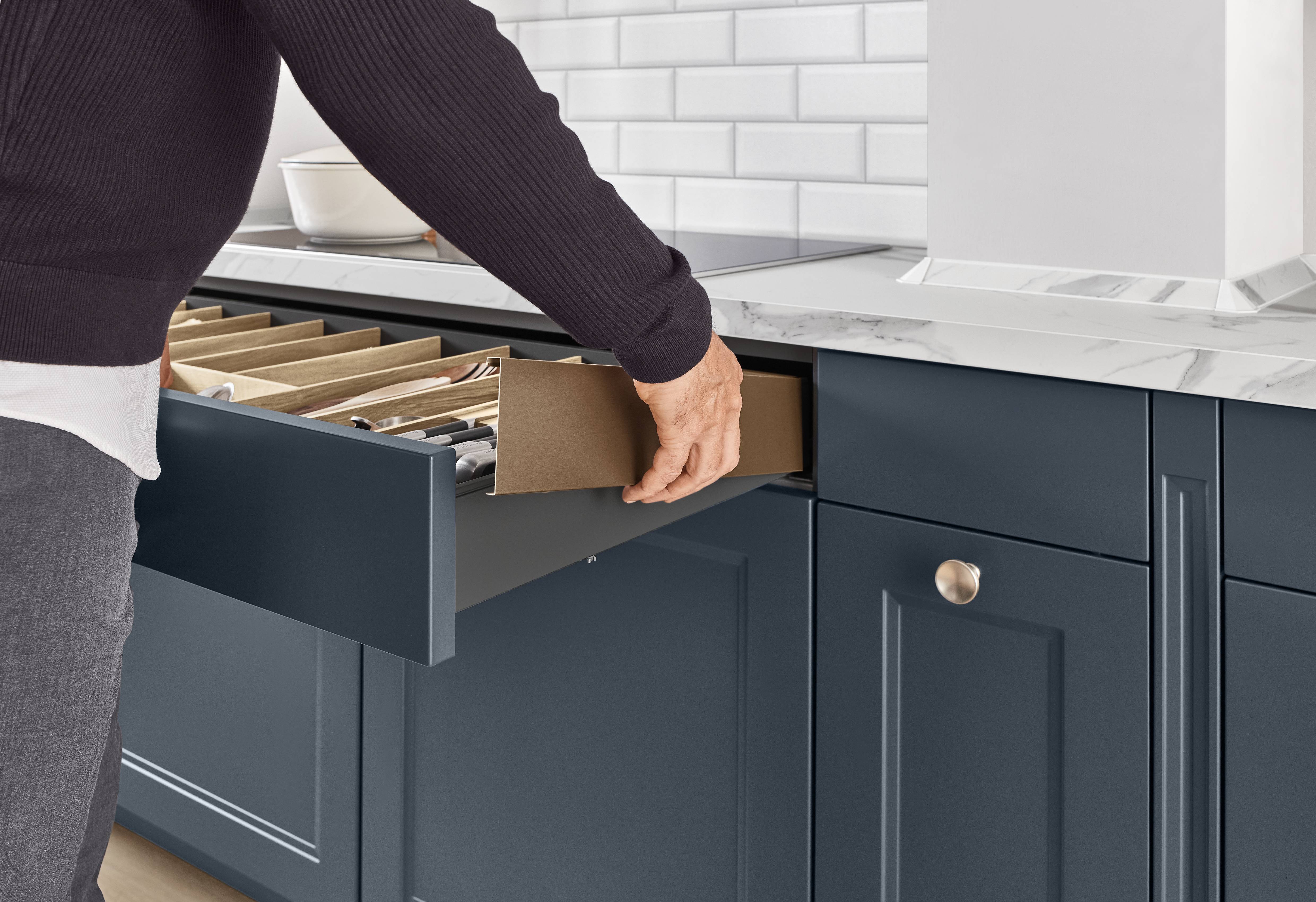 Cascada Kitchen — Navy Blue Cabinetry with Integrated Cutlery Drawer Organiser A person opening a wide deep drawer in navy blue Cascada kitchen cabinetry, revealing a wooden knife and cutlery organiser insert, with a marble worktop, white subway tile backsplash, and brushed nickel knob hardware visible.