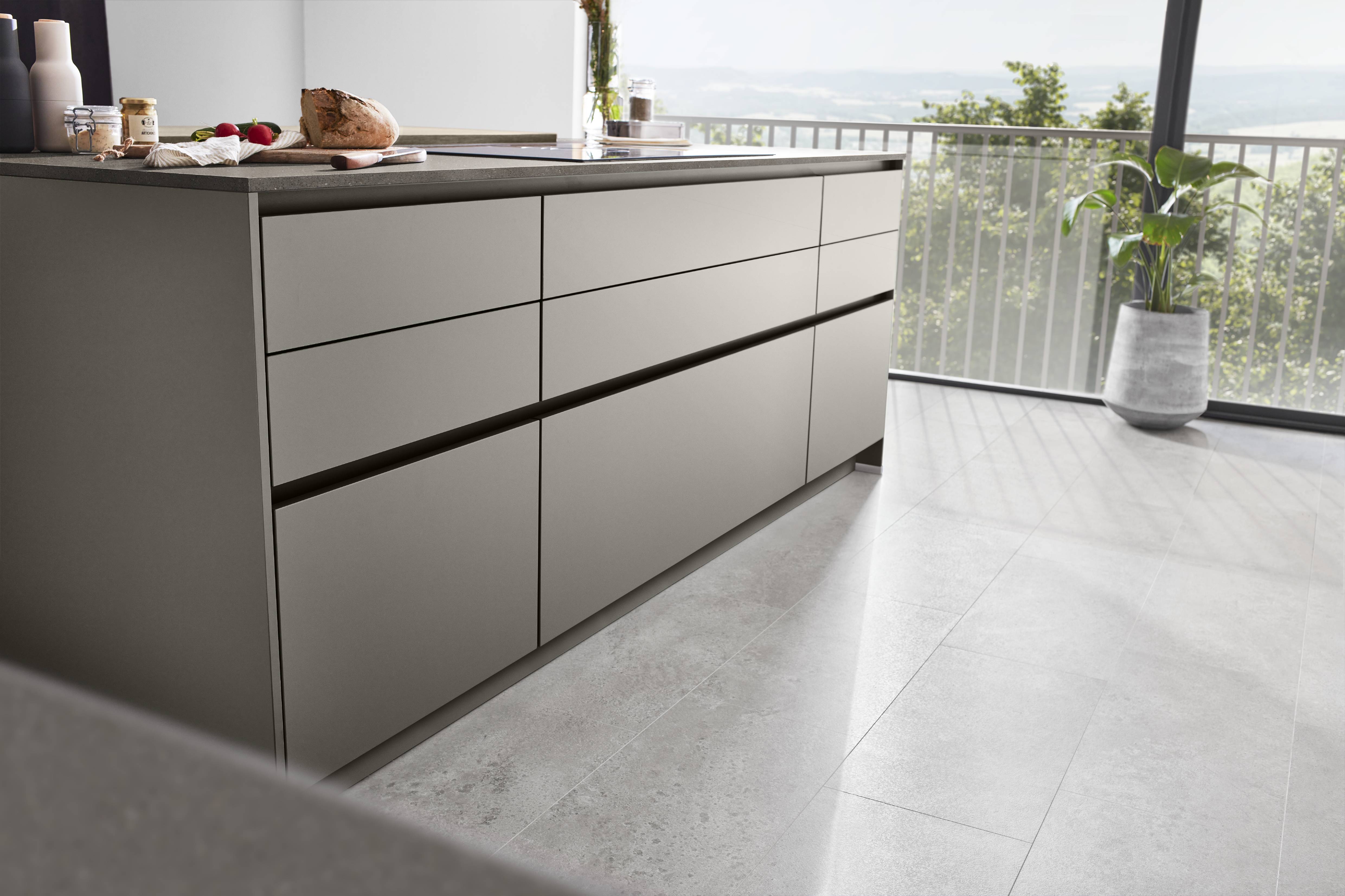 Riva Kitchen Island — Cashmere Grey with Stone Worktop Modern kitchen island with flat-front cashmere grey drawers, integrated dark bar handles, and a dark stone countertop, set against floor-to-ceiling windows with garden views.