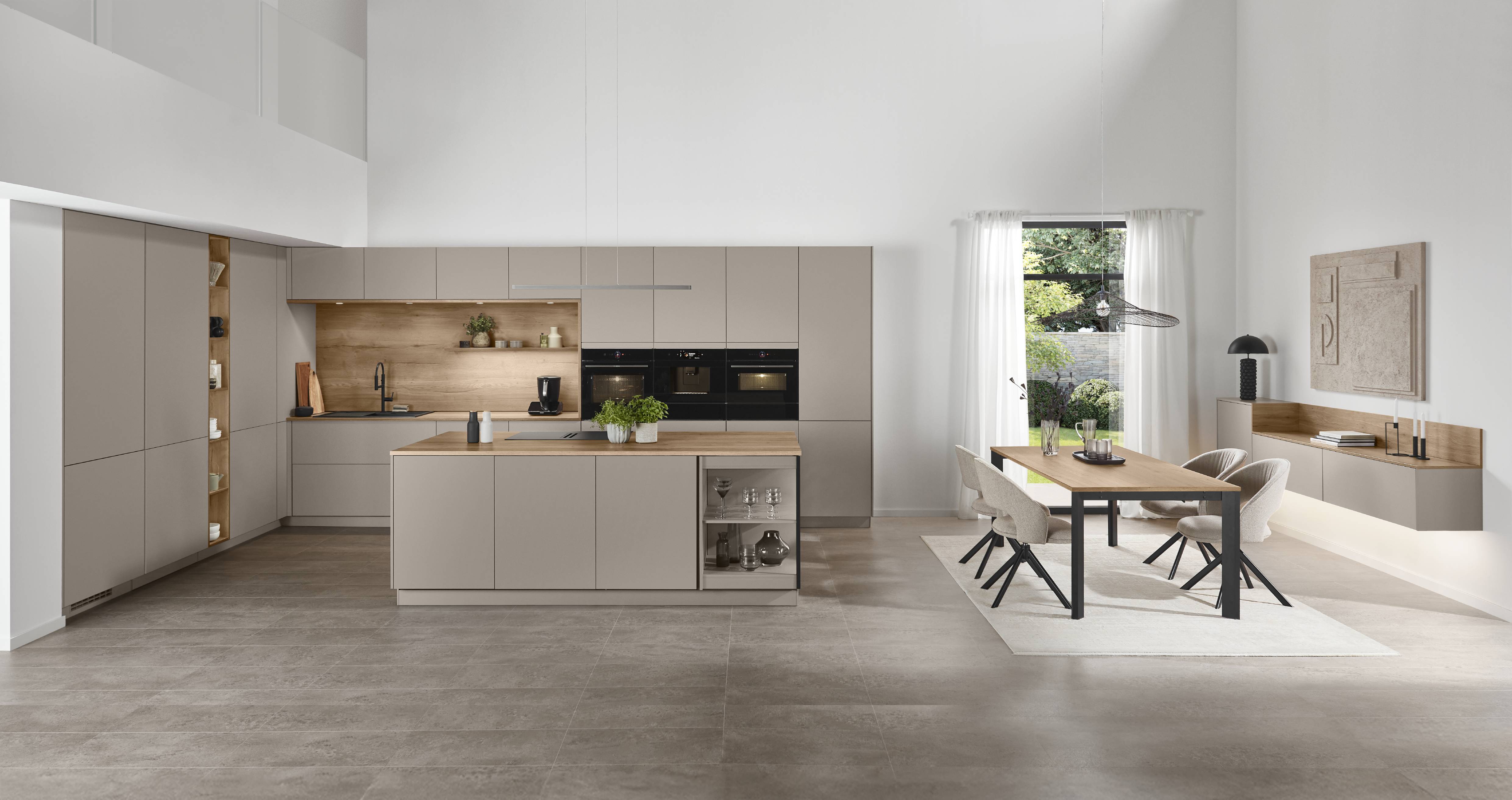 Open-plan Senso kitchen in greige with handleless cabinetry, oak splashback shelving with LED lighting, a central island with oak worktop and an open glass display niche, triple integrated black ovens, a tall oak-accented display cabinet, and a connected dining area with an oak table, upholstered chairs, and garden views through sheer curtains.