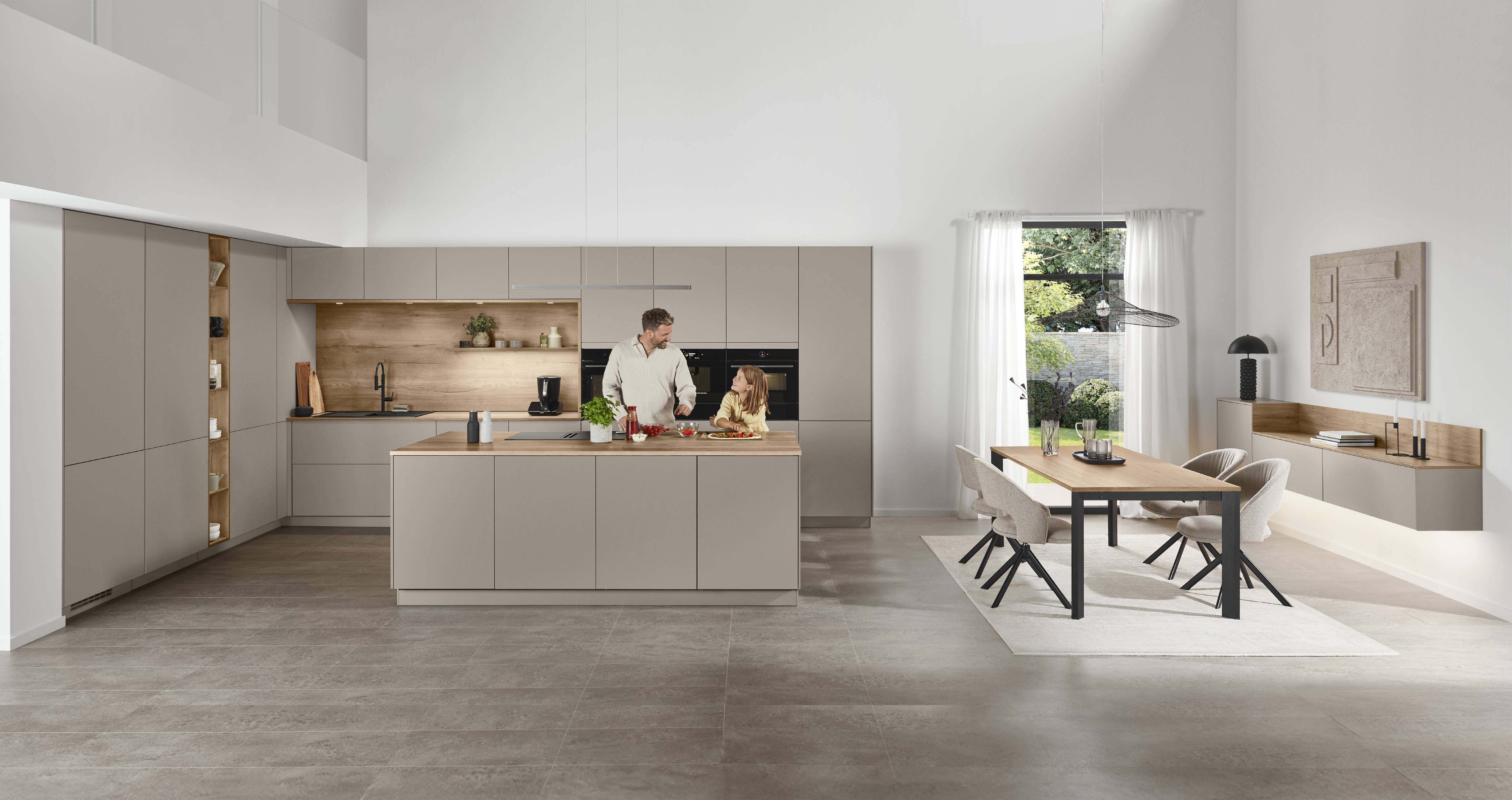 Senso Kitchen — Greige with Oak Accents, Open-Plan Living Spacious open-plan kitchen and dining area featuring handleless greige cabinetry, a central island with oak worktop, integrated black appliances, and a father and daughter cooking together.
