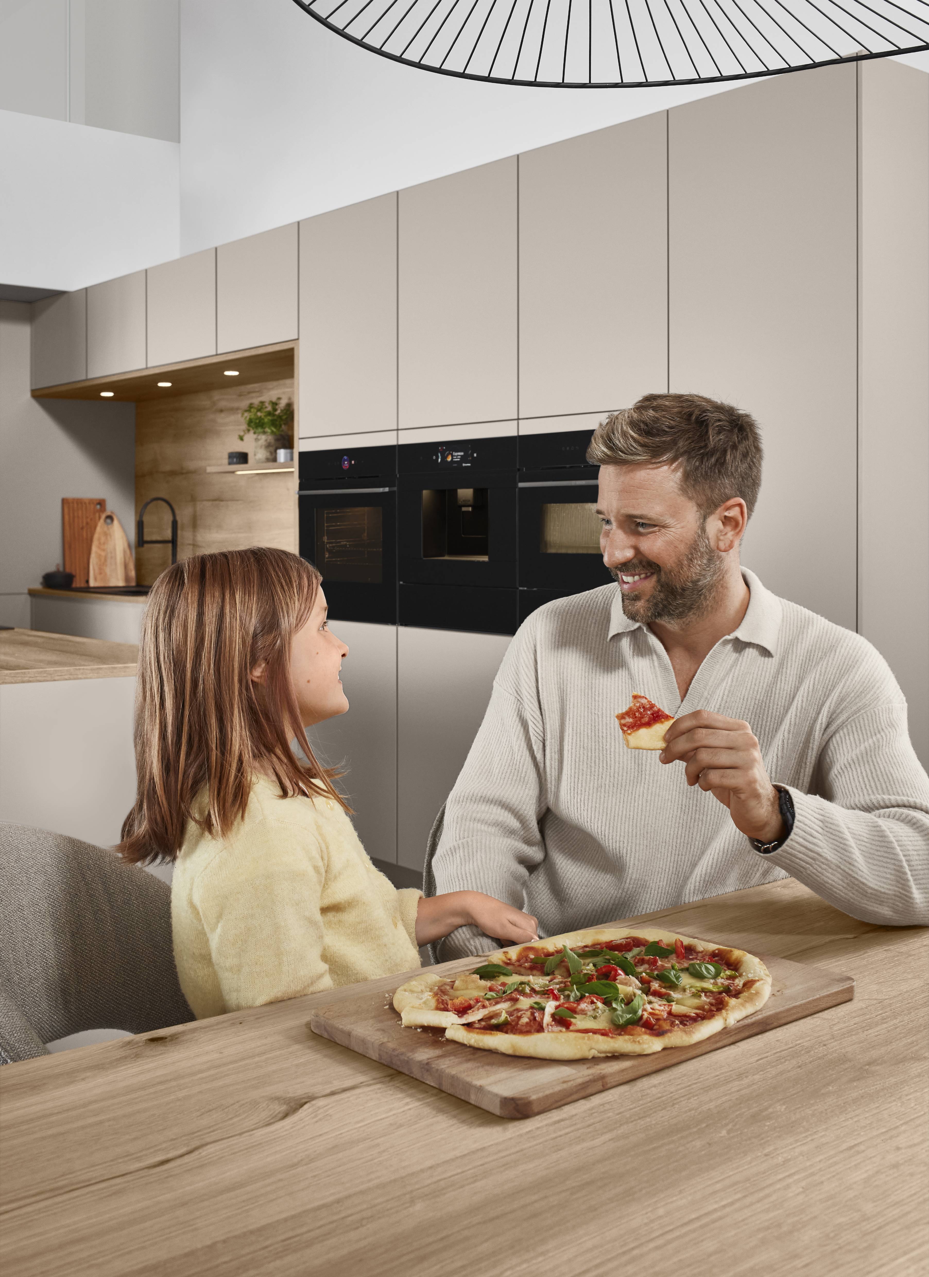 Senso Kitchen — Family Moments in Greige and Oak A smiling father and young daughter seated at a wooden kitchen island sharing a homemade pizza, with greige handleless cabinetry, oak shelving, and integrated black ovens visible in the background.