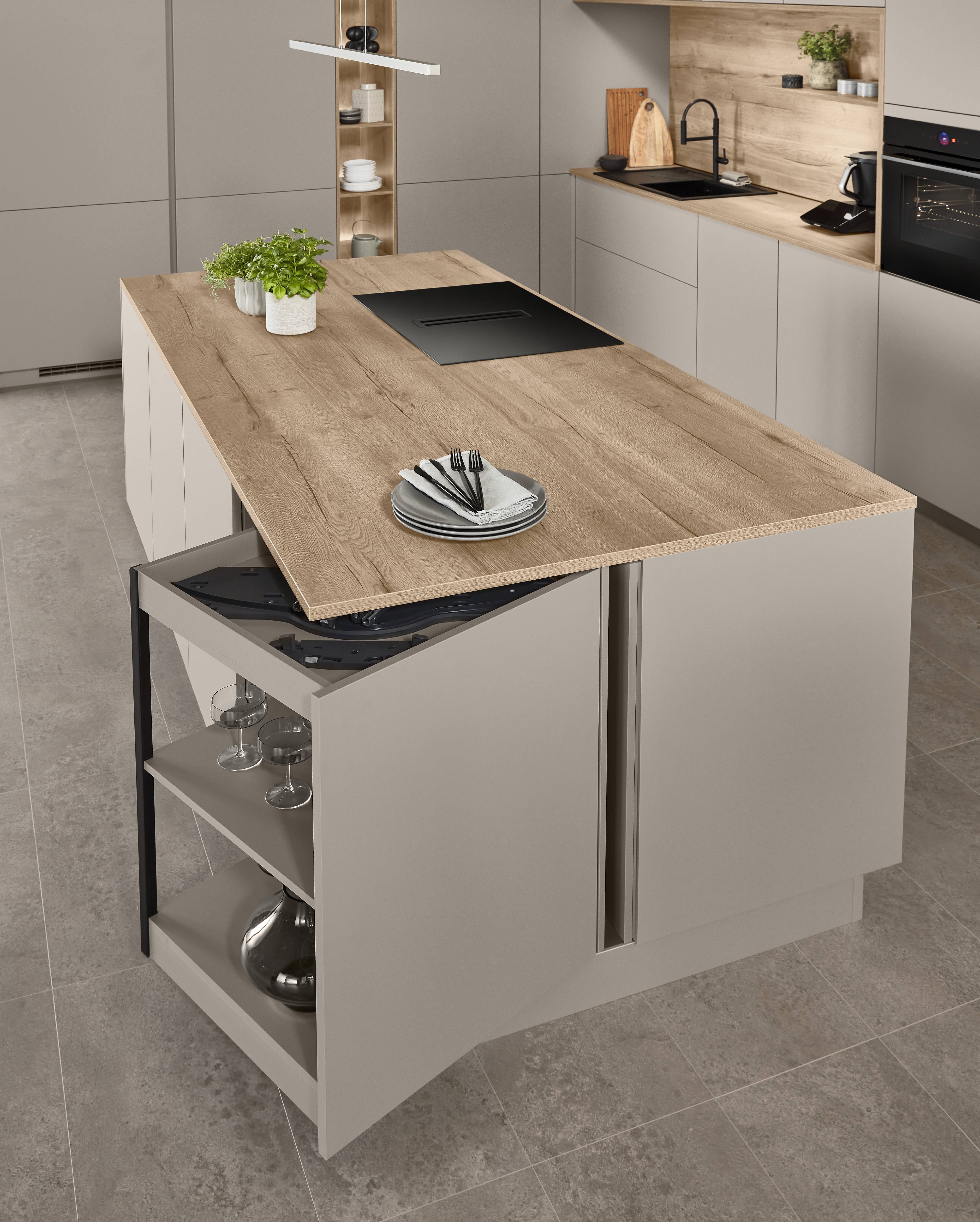 Senso Kitchen Island — Hidden Storage with Flip-Up Worktop Extension Aerial view of a Senso kitchen island in greige with an oak worktop, showing a lifted flip-up panel revealing concealed storage below, open side shelving with wine glasses, and a black induction hob on the surface.