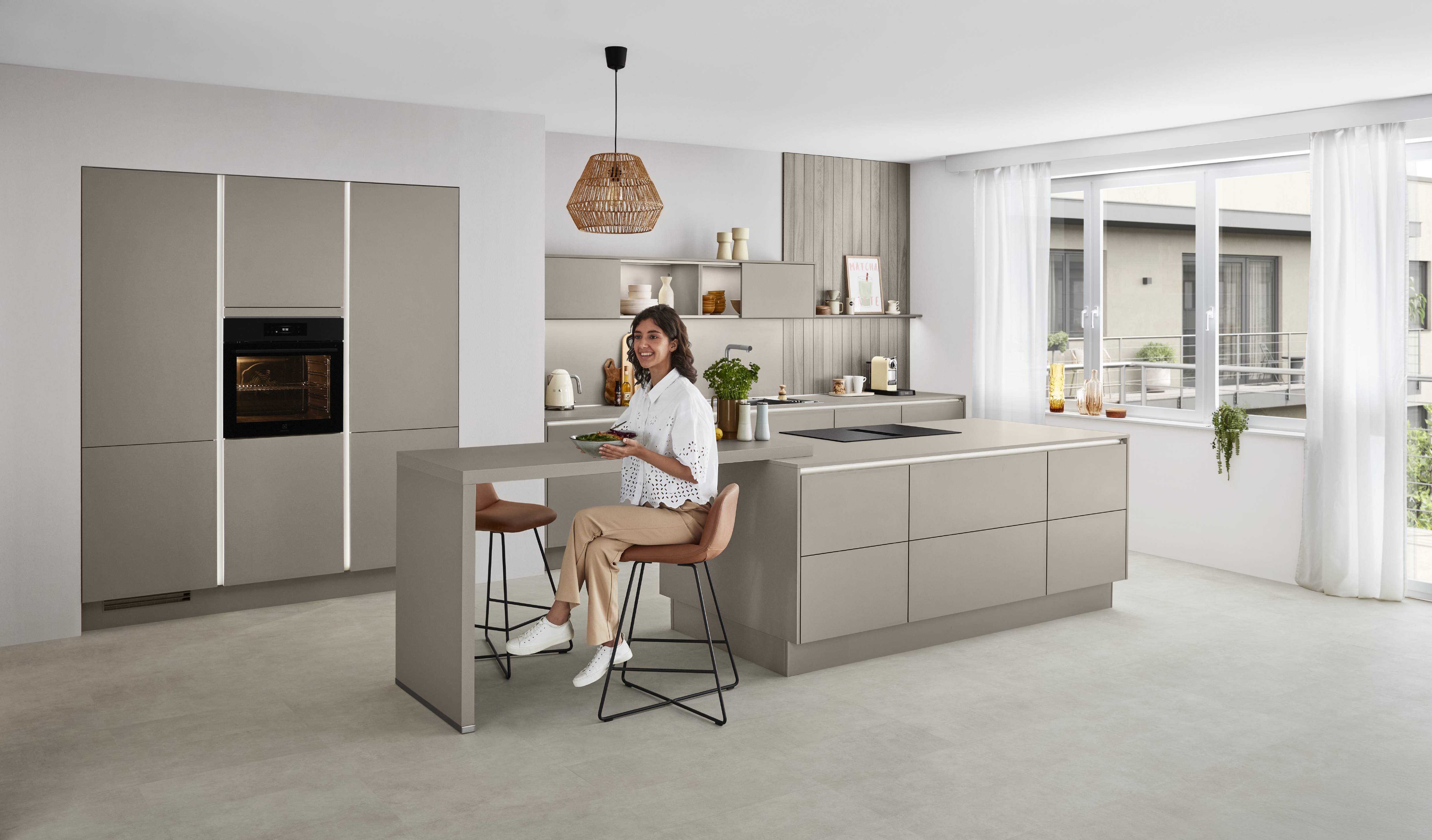 Laser Kitchen — Warm Taupe with Peninsula Bar and Integrated Lighting A smiling woman seated at a taupe kitchen peninsula bar holding a bowl, with a handleless Laser kitchen featuring open shelving, a tall cabinet with integrated lighting strip, black built-in oven, and balcony views behind sheer curtains.