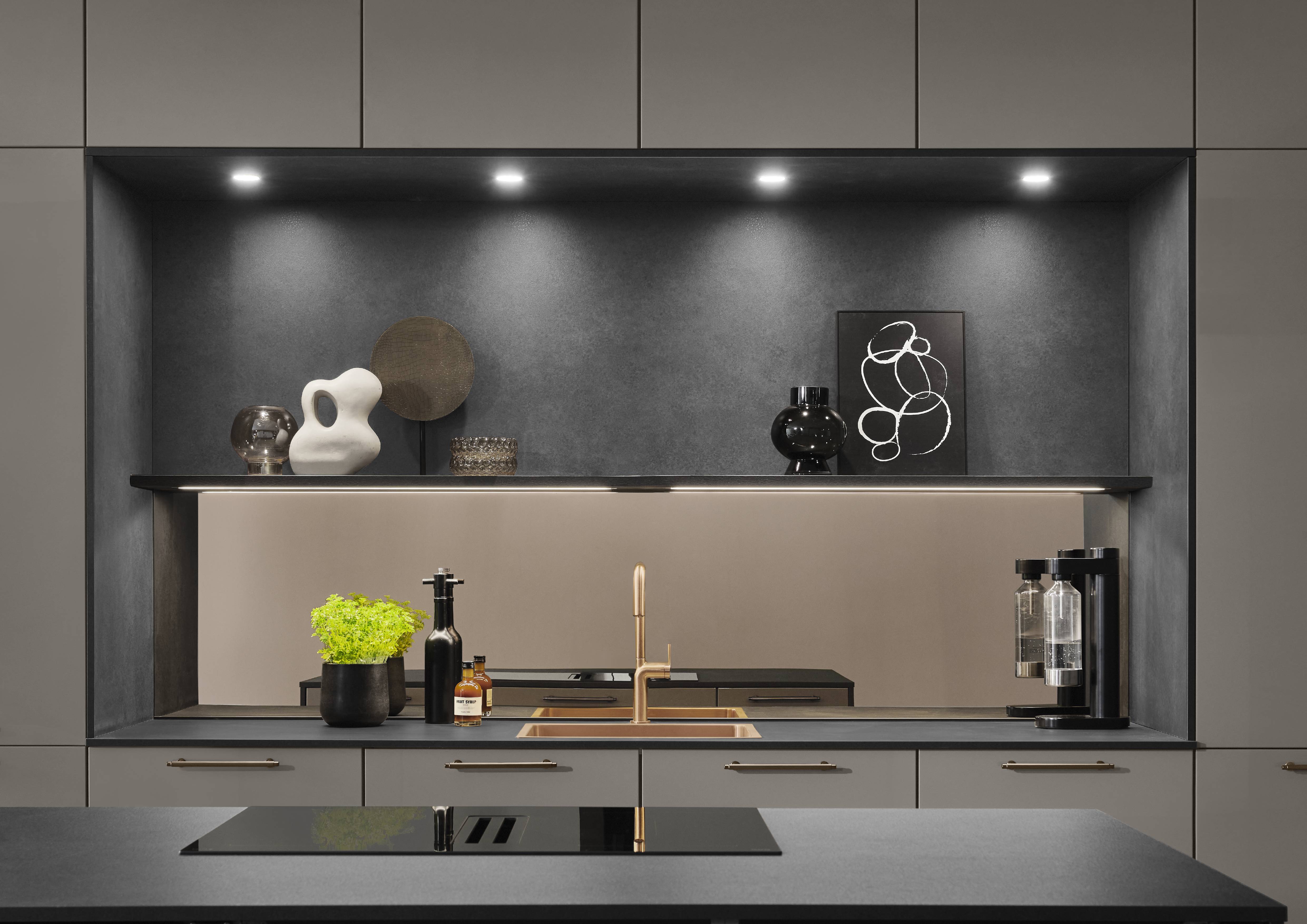 Close-up of a Noblessa kitchen cooking niche in greige with recessed ceiling spotlights, a black floating shelf displaying sculptural ceramics and abstract art, a bronze-tinted mirror backsplash, a rose gold undermount sink with a brushed gold mixer tap, brass bar handles on dark lower drawers, a black induction hob on the foreground island, and a herb plant and condiment bottles on the worktop.