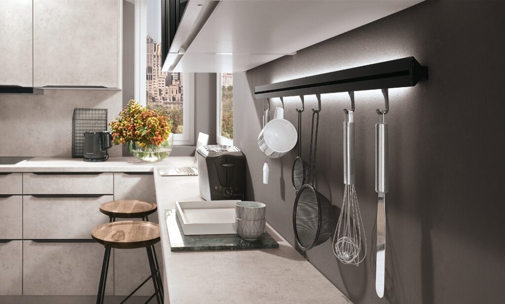 Close-up of a black wall-mounted LED-lit utensil rail holding hooks with a mug, strainer, and whisk, set in a concrete-effect grey kitchen with a white worktop, wooden stools, a coffee machine, a laptop, and a city view through the window.