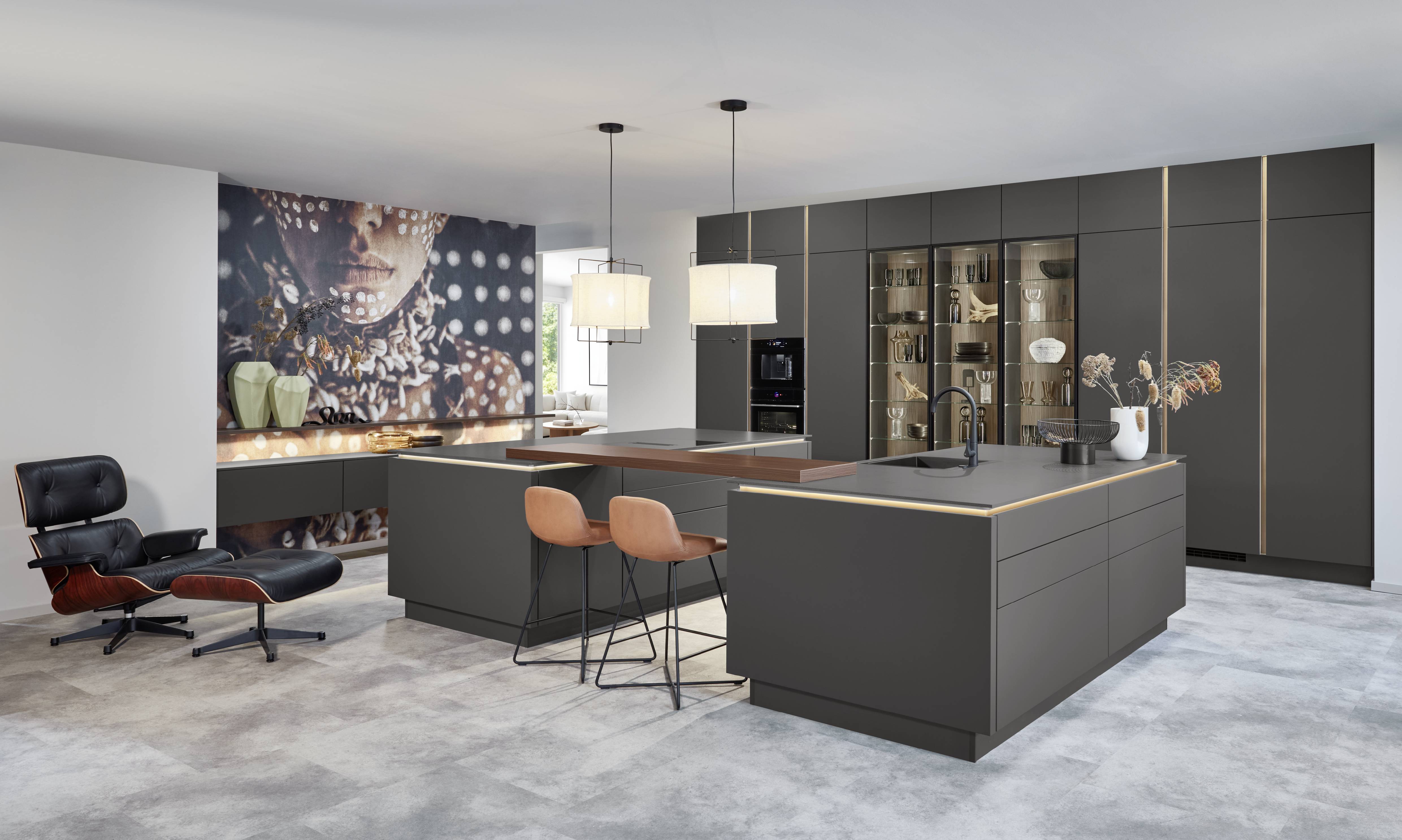 Graphite European Kitchen — Brass Trim, Glass Display Cabinet, and Dual LED Islands Luxury European kitchen with full-height matte graphite handleless cabinetry accented by vertical brass trim lines, two islands with LED-lit worktop edges and walnut bar extensions, cognac leather bar stools, an illuminated glass display cabinet with glassware and ceramics, integrated black oven, a large-format art print feature wall, and a classic Eames lounge chair.