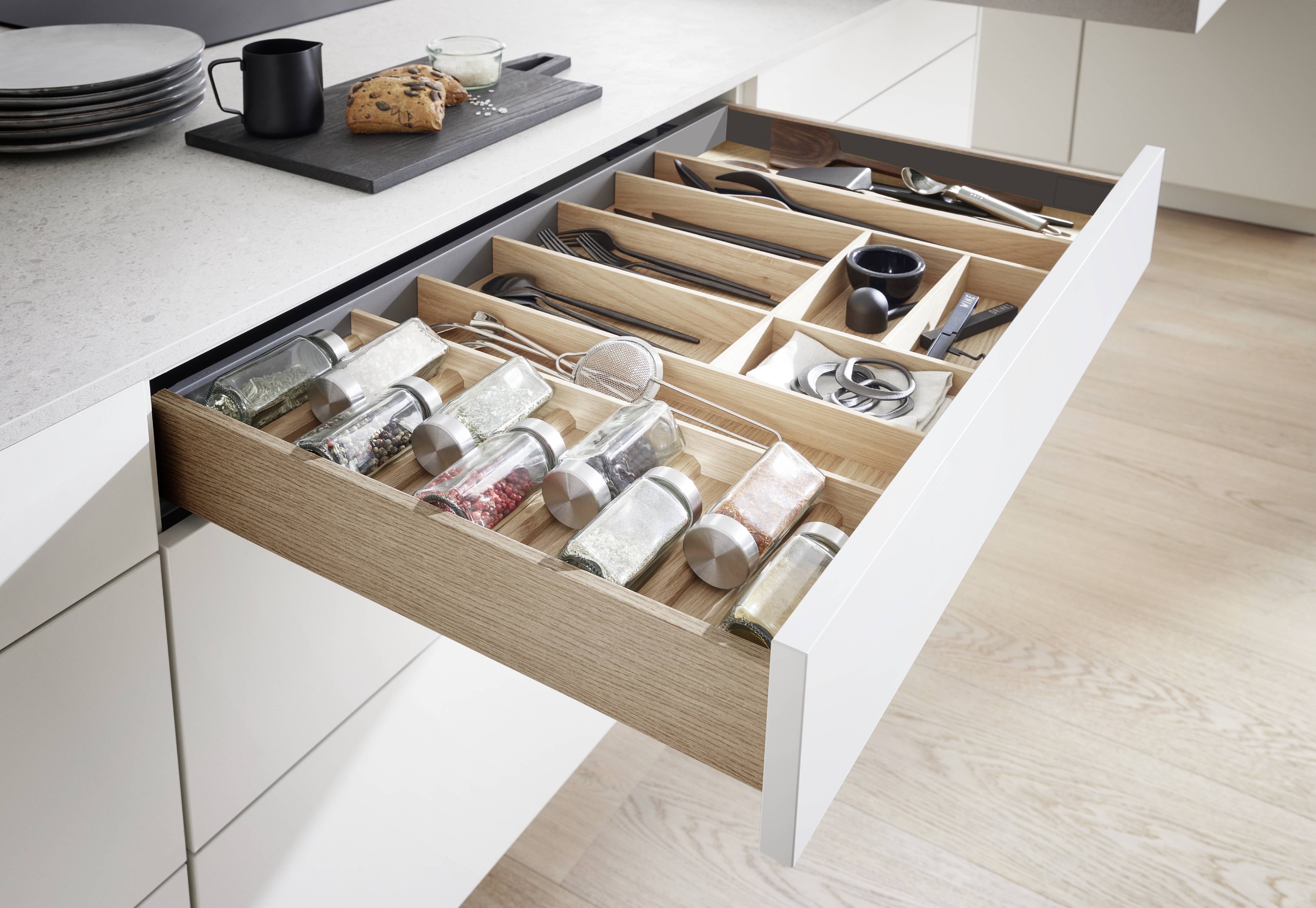 European Kitchen Drawer Organisation — Oak Divider Insert with Spice and Cutlery Storage Open wide kitchen drawer in white cabinetry revealing a fitted oak wood divider insert with two tiers, organising spice jars with stainless steel lids in the lower section and black cutlery, utensils, and small accessories in the upper divided compartments, on a white quartz worktop with a dark serving board above.