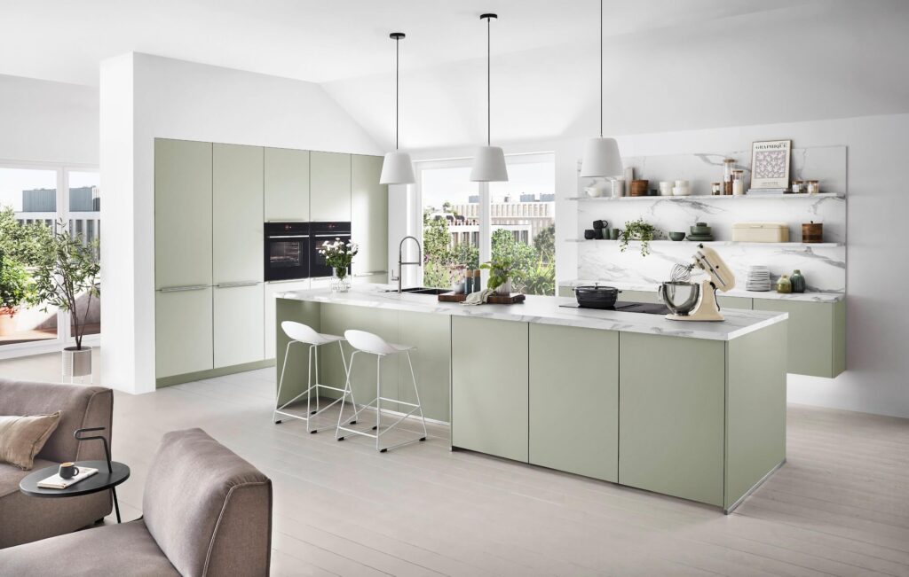 Modular done beautifully — sage green cabinetry, an oversized island, and open marble-backed shelving show what thoughtful kitchen remodeling can achieve.