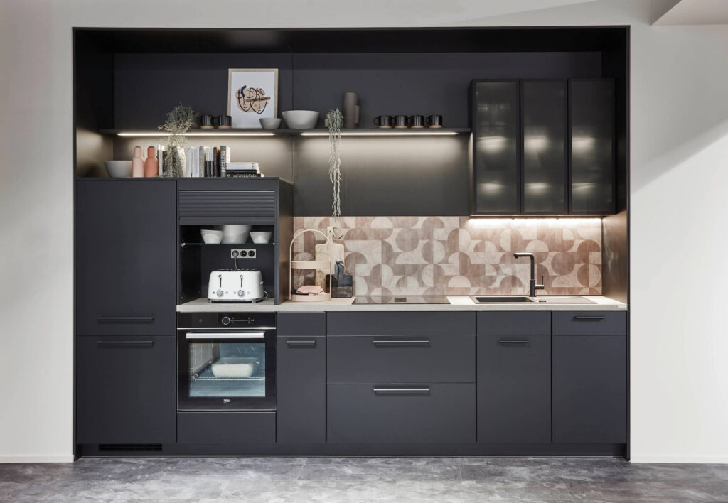 Compact matte charcoal kitchen in a recessed alcove, featuring frosted glass upper cabinets, open shelving with decor and books, integrated black oven and roller-shutter appliance garage, a black tap, and a patterned geometric backsplash.