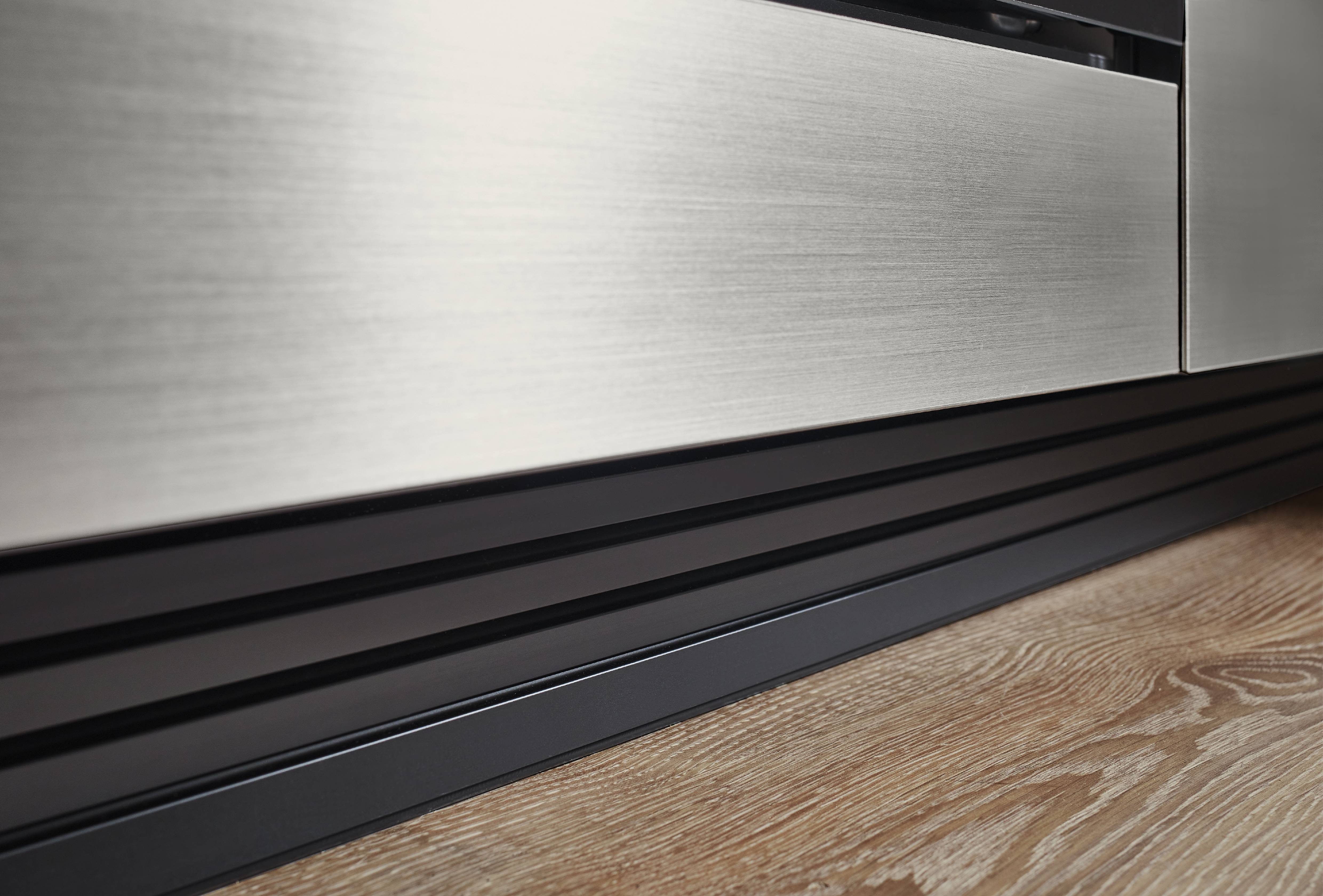 Brushed Stainless Steel Cabinet Front with Black Grooved Plinth — German Kitchen Finish Detail Extreme close-up product detail of a brushed stainless steel kitchen cabinet drawer front with a matte black multi-channel grooved aluminium plinth at the base, set against a light oak wood floor, showing precise German manufacturing quality and material contrast.