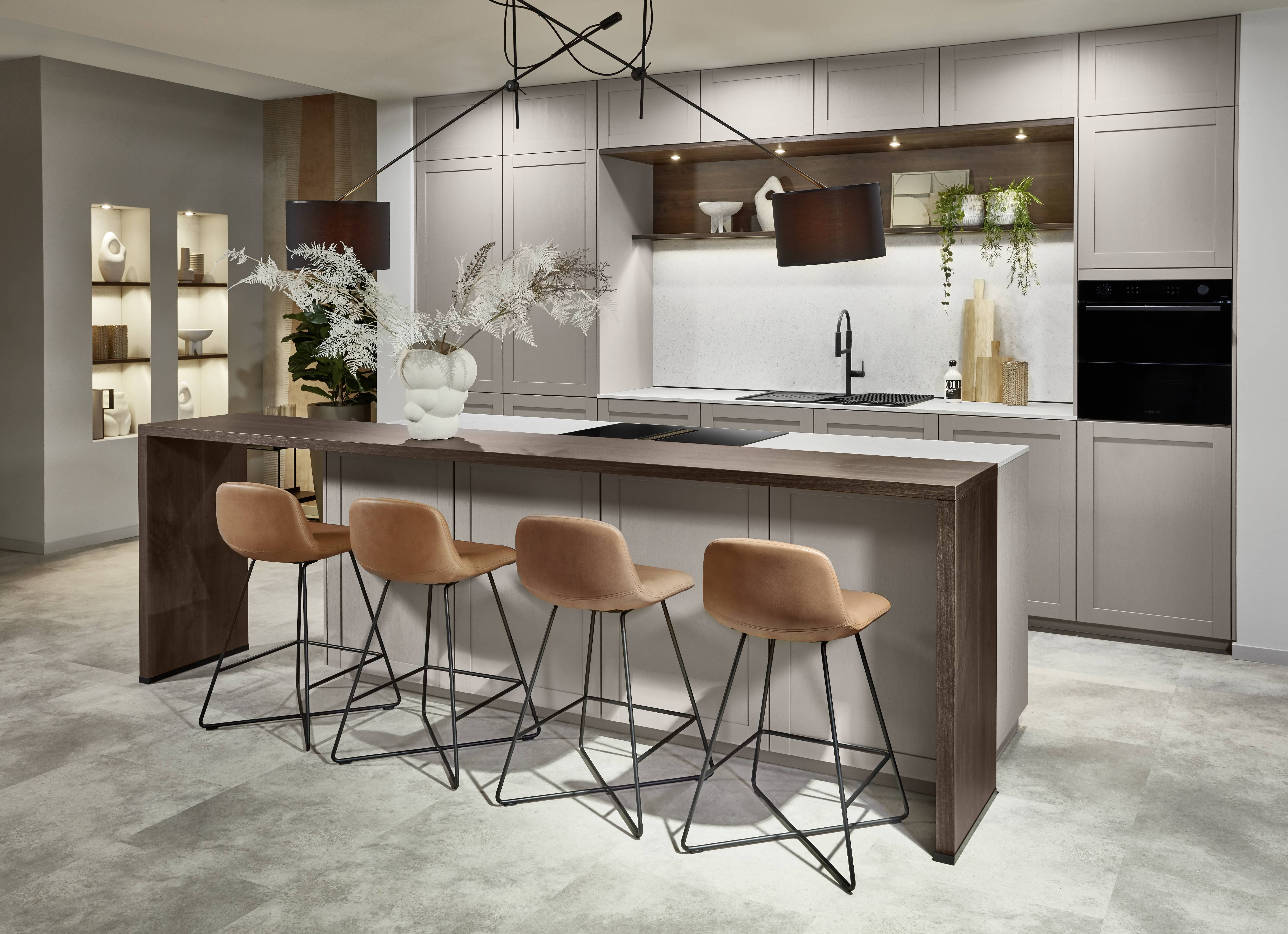 Greige Shaker Kitchen with Dark Walnut Island Bar — Classic Cabinet Style for Modern Spaces Modern shaker-style kitchen with greige frame-and-panel cabinetry, an integrated black hob and white worktop, a dark walnut island bar extension with three cognac velvet and black metal bar stools, an abstract black pendant light, illuminated display niches with sculptural ceramics, an oak open shelf with LED lighting, black sink and tap, integrated black oven, and concrete-effect floor tiles.