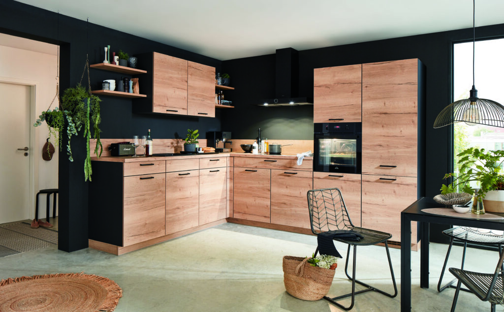 Rustic Oak Kitchen with Black Walls — Bold High-End Cabinet Design L-shaped kitchen with light rustic oak cabinetry and matching worktop, matte black walls, black bar handles, integrated black oven, open floating shelves with plants and ceramics, wire dining chairs, and trailing indoor plants throughout.