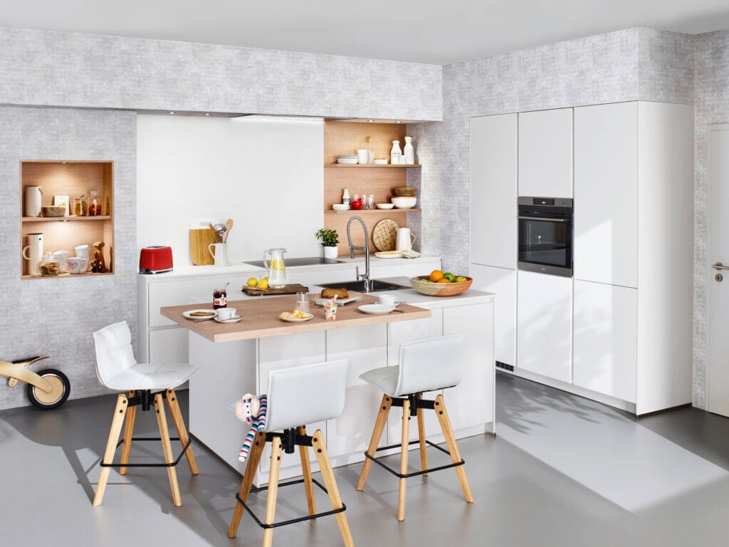 White Gloss Kitchen with Oak Shelving — High-End Cabinetry for Family Living Modern white gloss handleless kitchen with a central island and oak breakfast bar extension, Scandinavian-style white bar stools with wooden legs, illuminated oak wall niches, open shelving, and an integrated black oven in a tall cabinet.