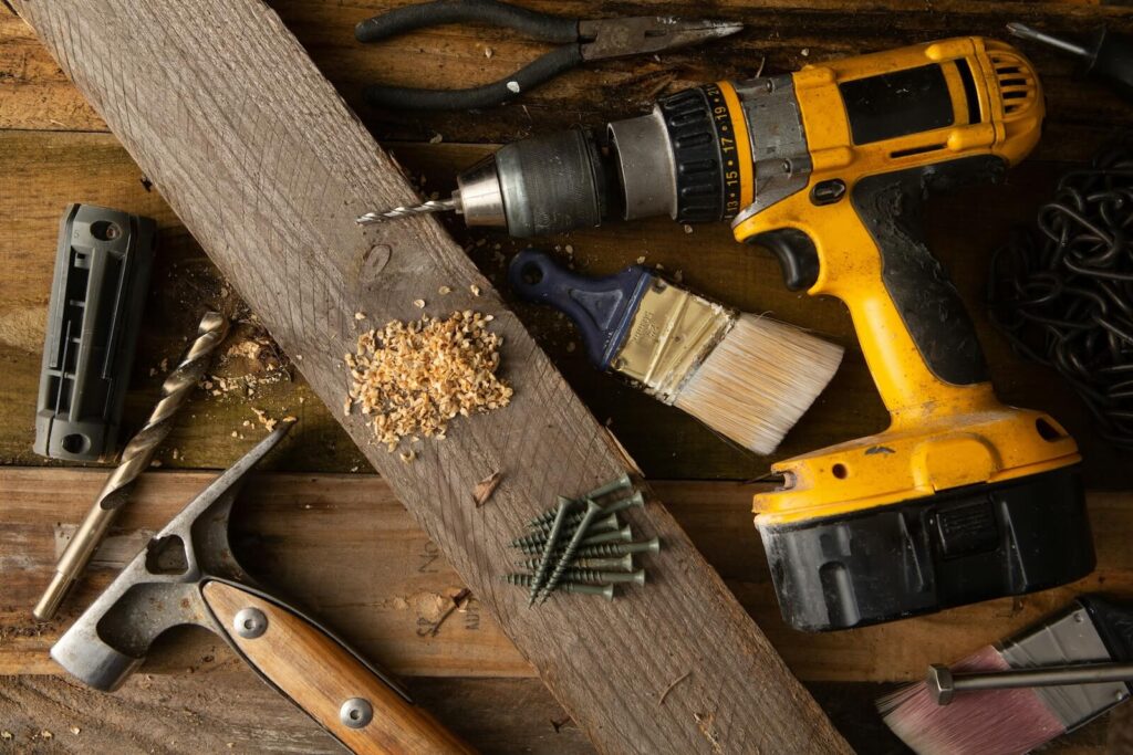 Overhead flat-lay photo of renovation tools on a wooden workbench, including a yellow power drill boring into a wood plank, a hammer, pliers, a paintbrush, wood screws, and sawdust shavings.