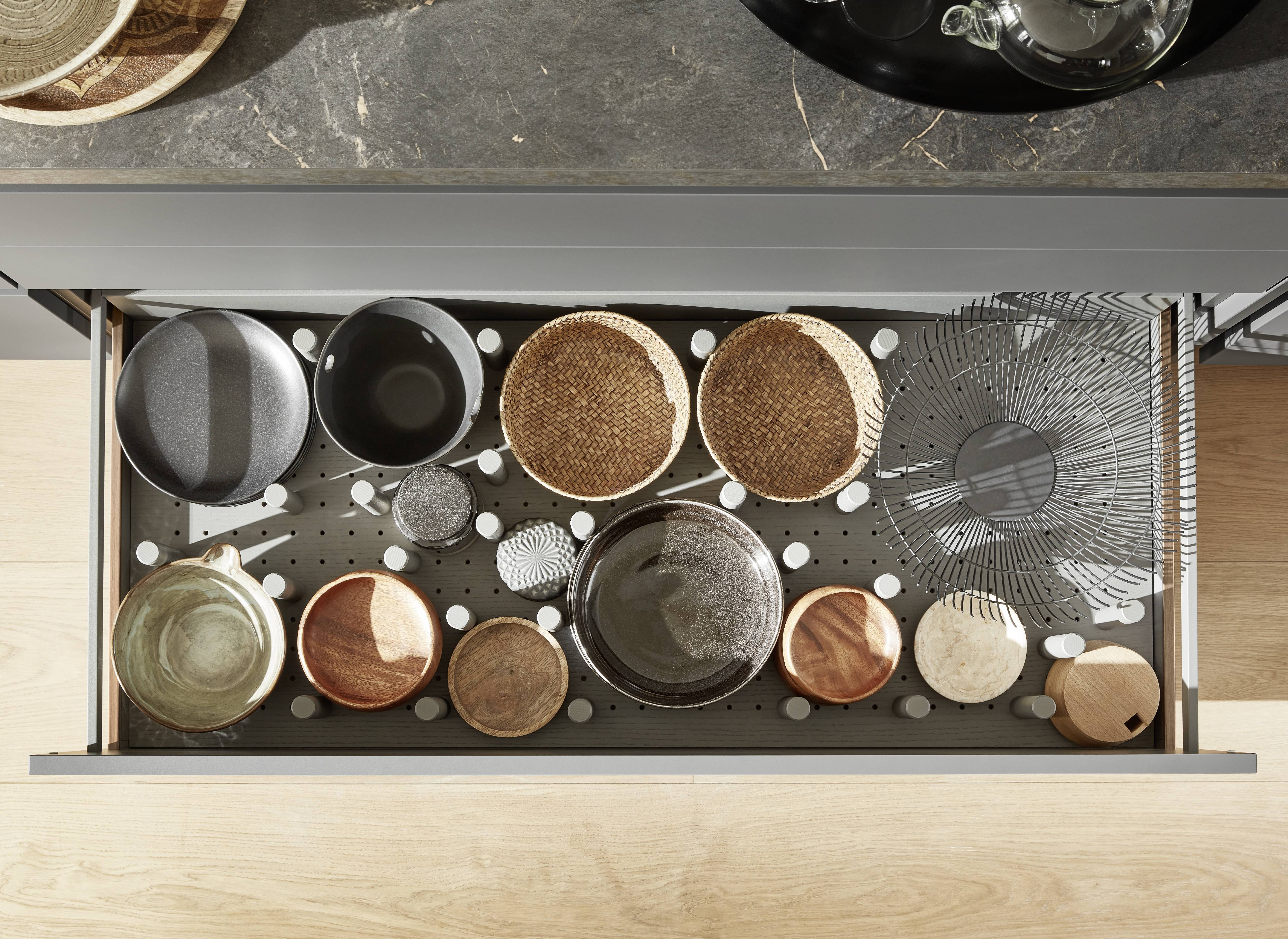 Deep Kitchen Drawer with Pegboard Base — Flexible Cookware and Bowl Storage Solution Top-down view of a deep open kitchen drawer with a dark pegboard base fitted with white adjustable pegs, storing a grey non-stick pan, black cookware, two woven rattan baskets, a wire serving dish, assorted ceramic and wooden bowls, and small decorative accessories, beneath a dark marble-effect stone worktop.