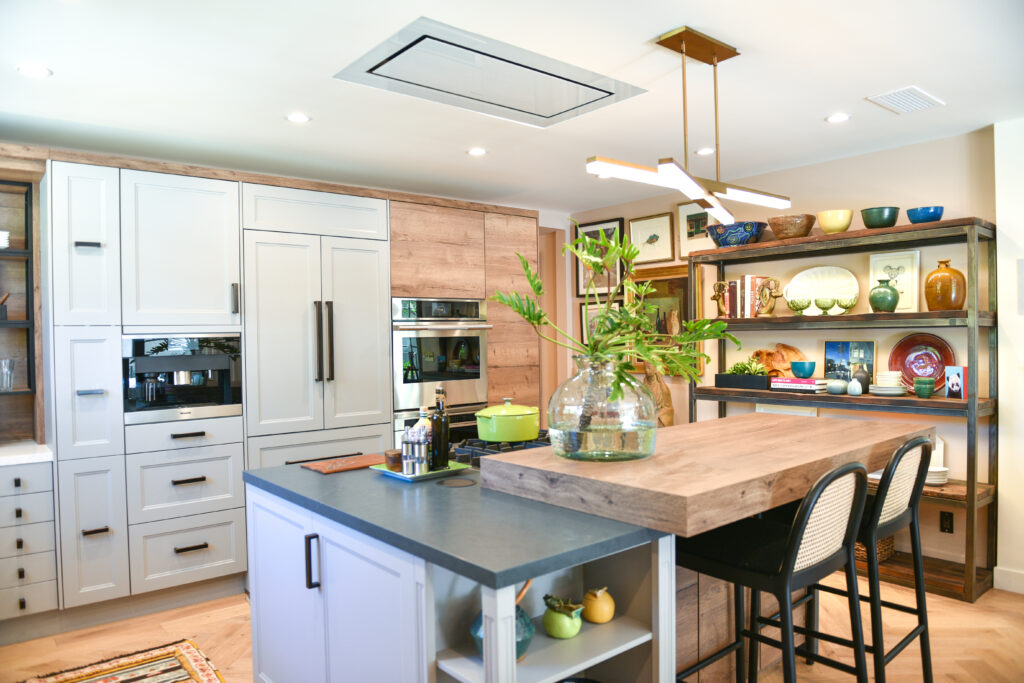 Transitional kitchen with off-white shaker-style cabinetry, a two-tone island with grey worktop and butcher-block wood extension, ceiling-flush range hood, stainless steel built-in ovens, industrial shelving unit with colourful ceramics and books, and a brass linear pendant light above.