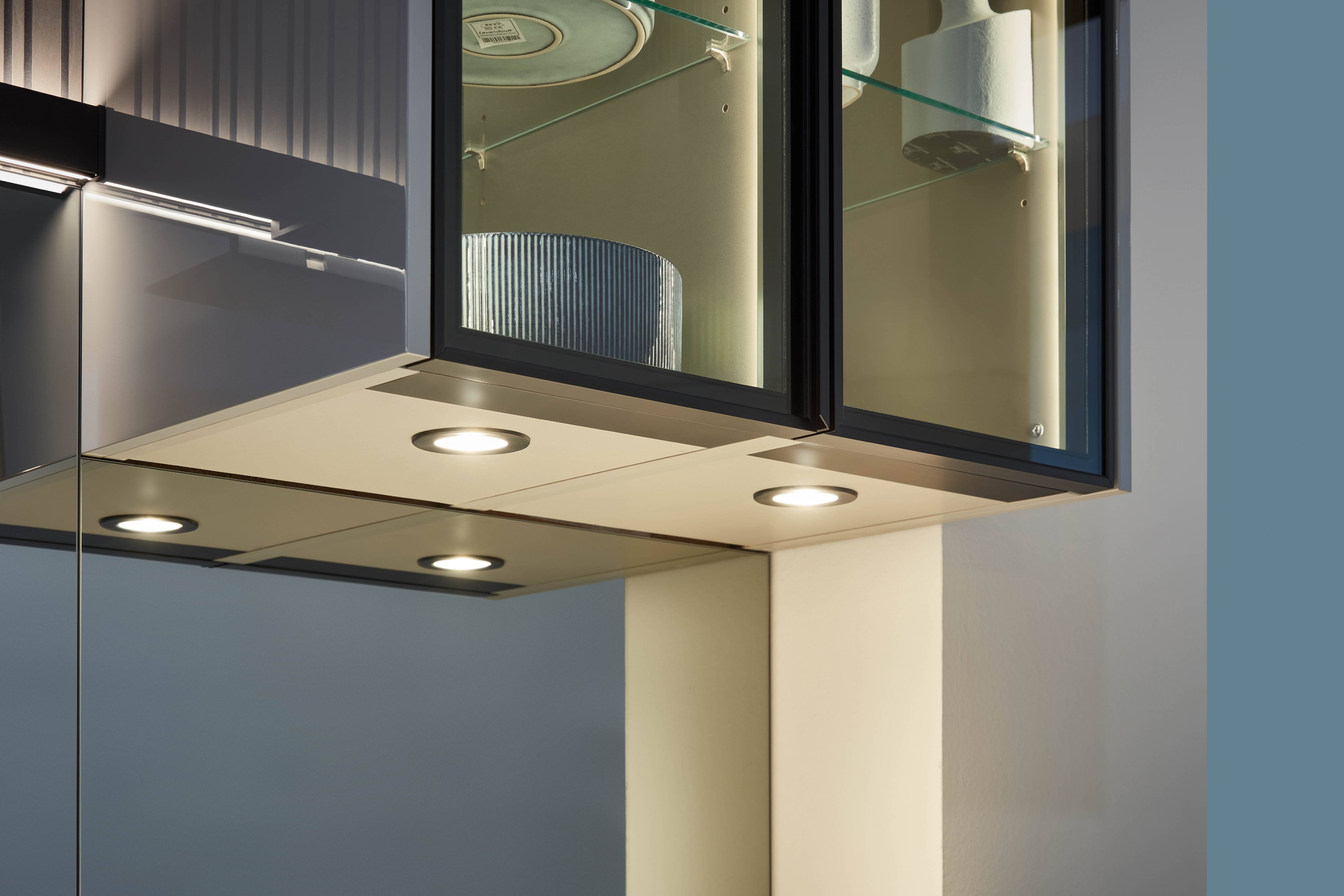 Kitchen Under-Cabinet LED Downlights — Integrated Lighting for Glass Display Cabinets Close-up detail of kitchen under-cabinet lighting showing three recessed LED downlights mounted on a cream-toned cabinet base, an open glass-fronted display cabinet with warm internal lighting revealing crockery on glass shelves, dark framing, and a mirrored cabinet surface reflecting the setup.