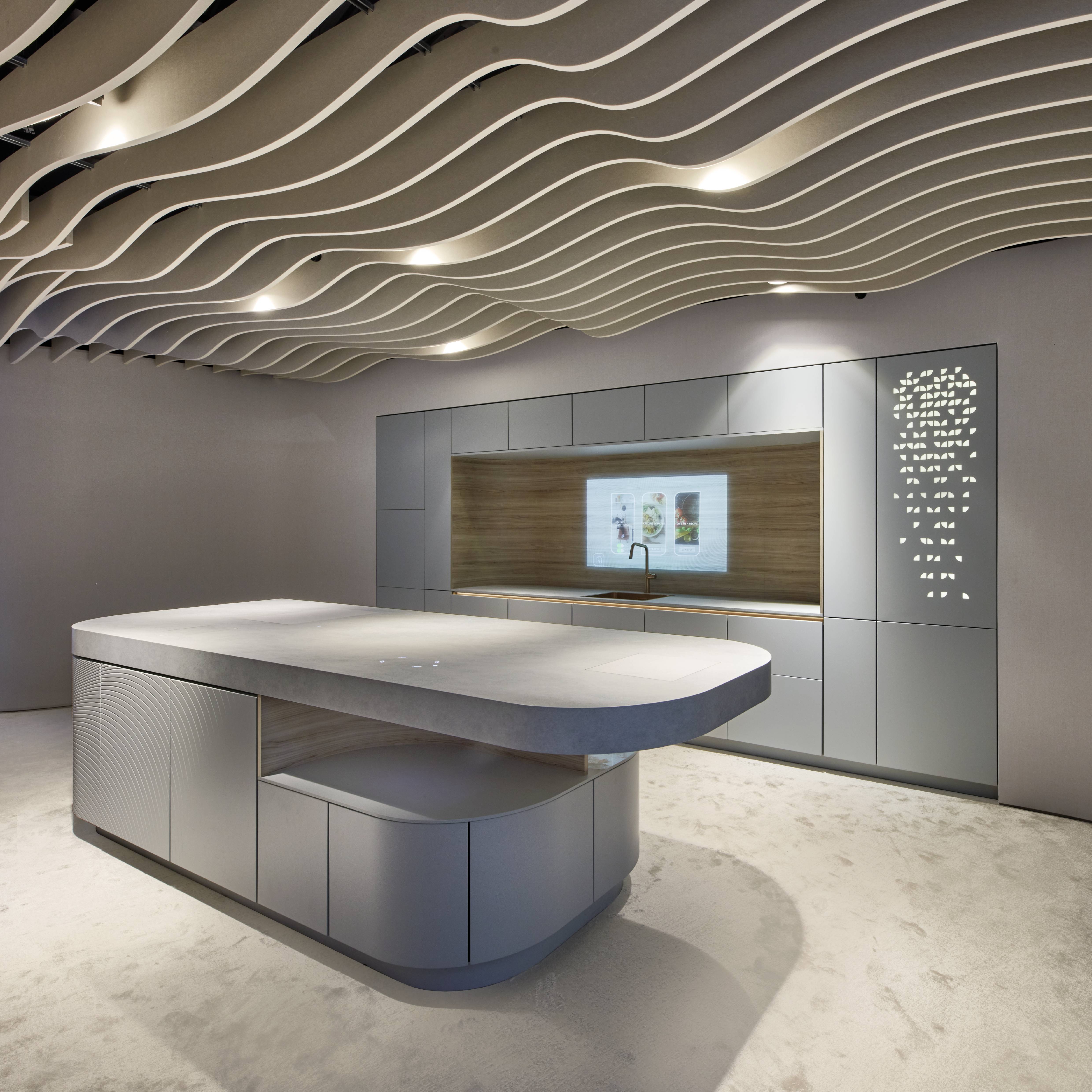 Futuristic Curved Kitchen Exhibition Display — Smart Screen, Arabic Laser-Cut Panel, and Wave Ceiling High-end kitchen exhibition display featuring a large curved grey island with a white stone worktop and sculpted concentric relief panels on its base, a grey handleless wall cabinet run with an integrated smart screen showing food content, an Arabic geometric laser-cut white decorative panel, a dramatic undulating wave ceiling installation with recessed spotlights, and a polished concrete floor.