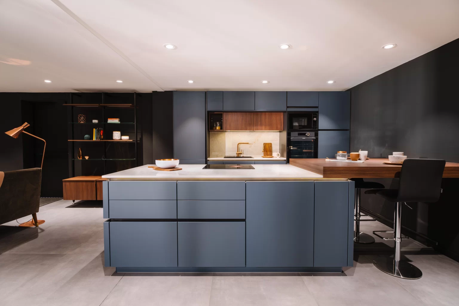 Contemporary kitchen with a large slate blue handleless island featuring a white marble worktop, brass edge detail, and integrated hob, connected to a walnut dining bar with black bar stools, full-height matte blue cabinetry flanking a walnut and brass appliance niche with gold tap, and a black open shelving unit in the background.