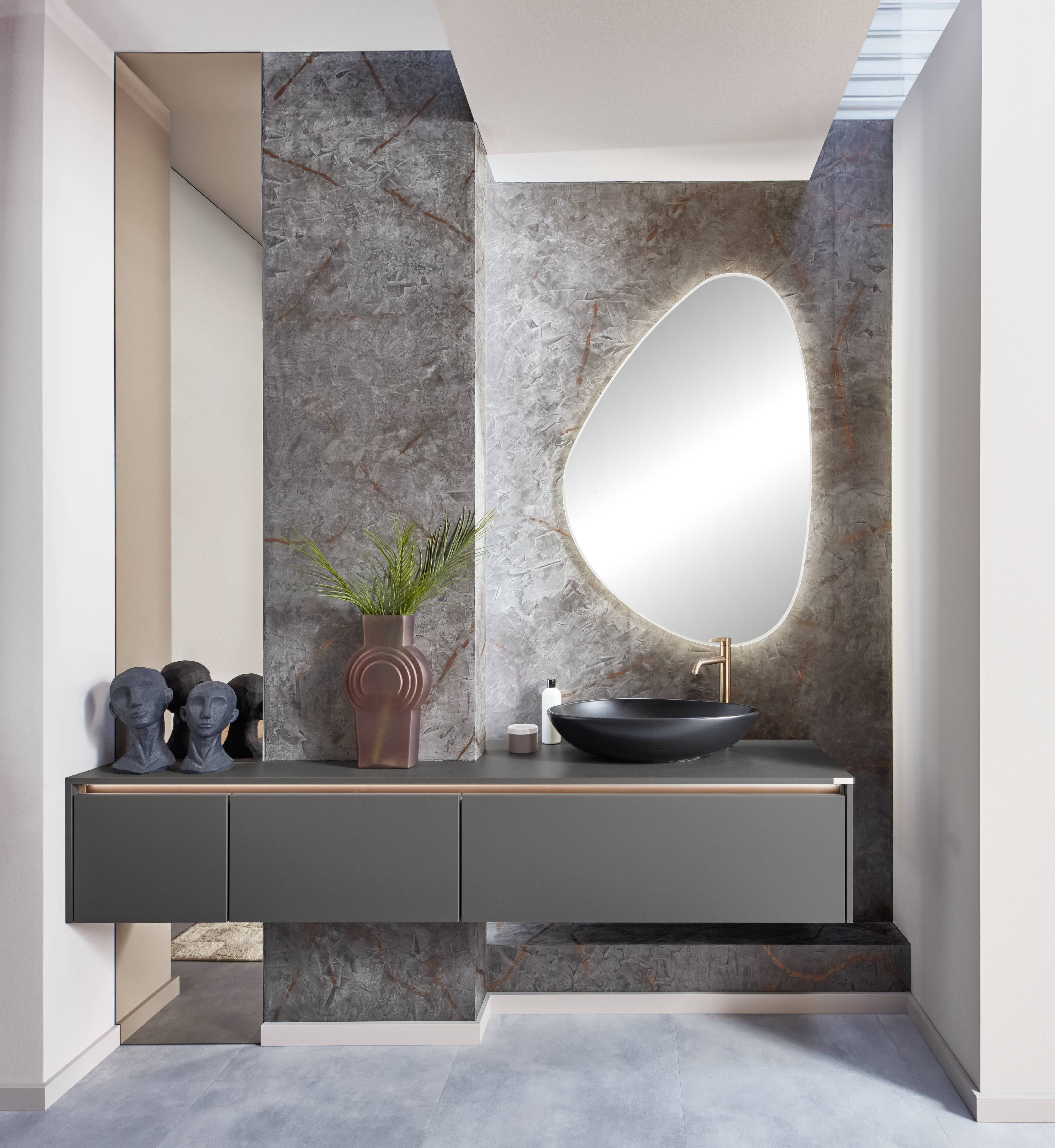 Contemporary Olive Bathroom Vanity — Asymmetric Mirror, Stone Wall, and Brushed Gold Fixtures Contemporary floating bathroom vanity in matte olive grey with three drawers, a gold-toned integrated handle rail, a dark stone worktop, a matte black oval vessel sink, a brushed gold mixer tap, an asymmetric freeform backlit mirror, a dramatic grey and rust-veined stone feature wall, sculptural ceramic head decorative objects, a terracotta vase with palm leaf, and grey floor tiles.