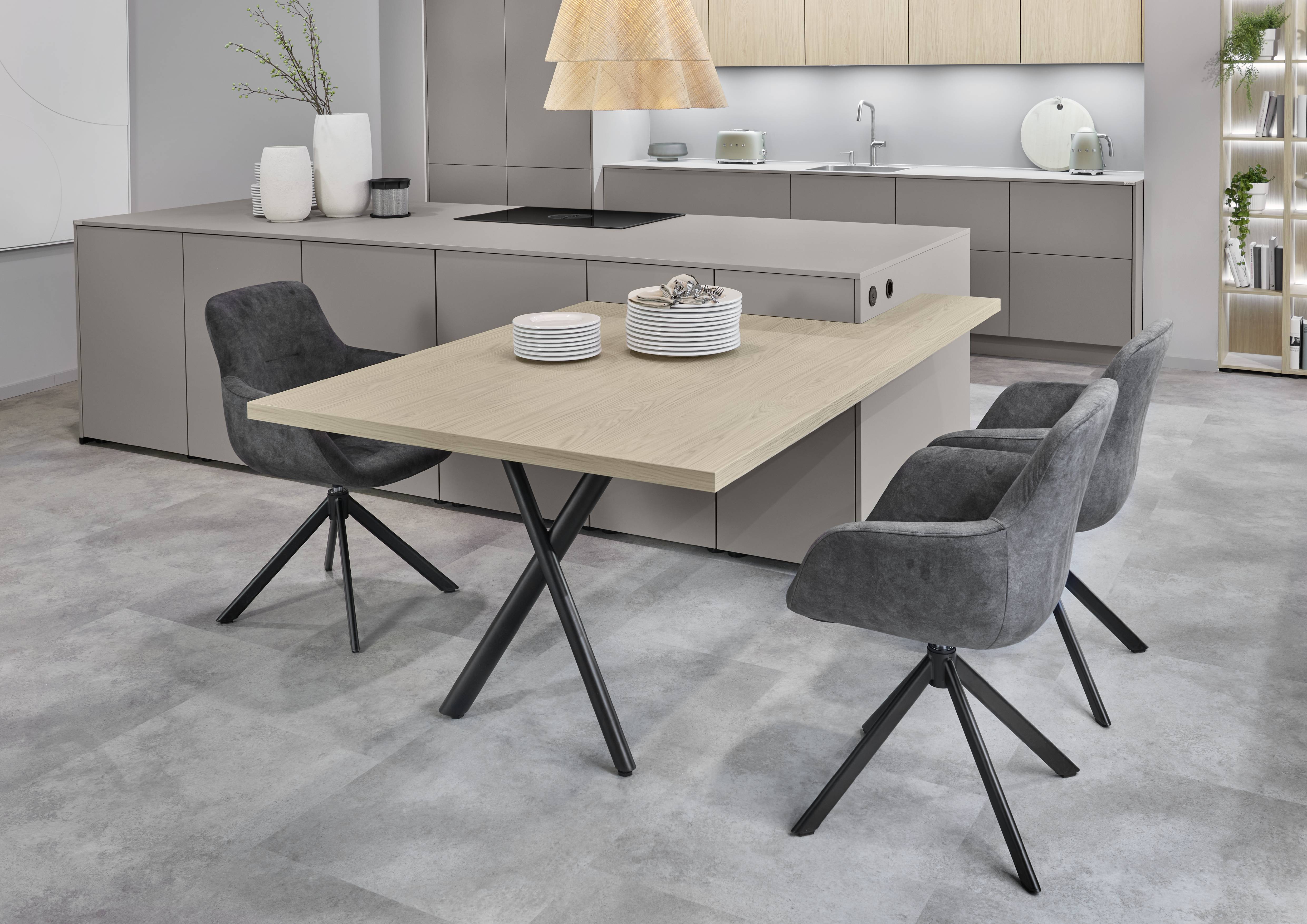 Greige Kitchen with Oak Dining Extension — Where Modern Meets Contemporary Design Contemporary kitchen and dining space with a greige handleless island featuring an integrated induction hob, a connected oak dining table with black star-base legs, dark grey upholstered swivel dining chairs, greige upper cabinetry with oak accents, a woven rattan pendant light, an illuminated glass display cabinet, and concrete-effect floor tiles.