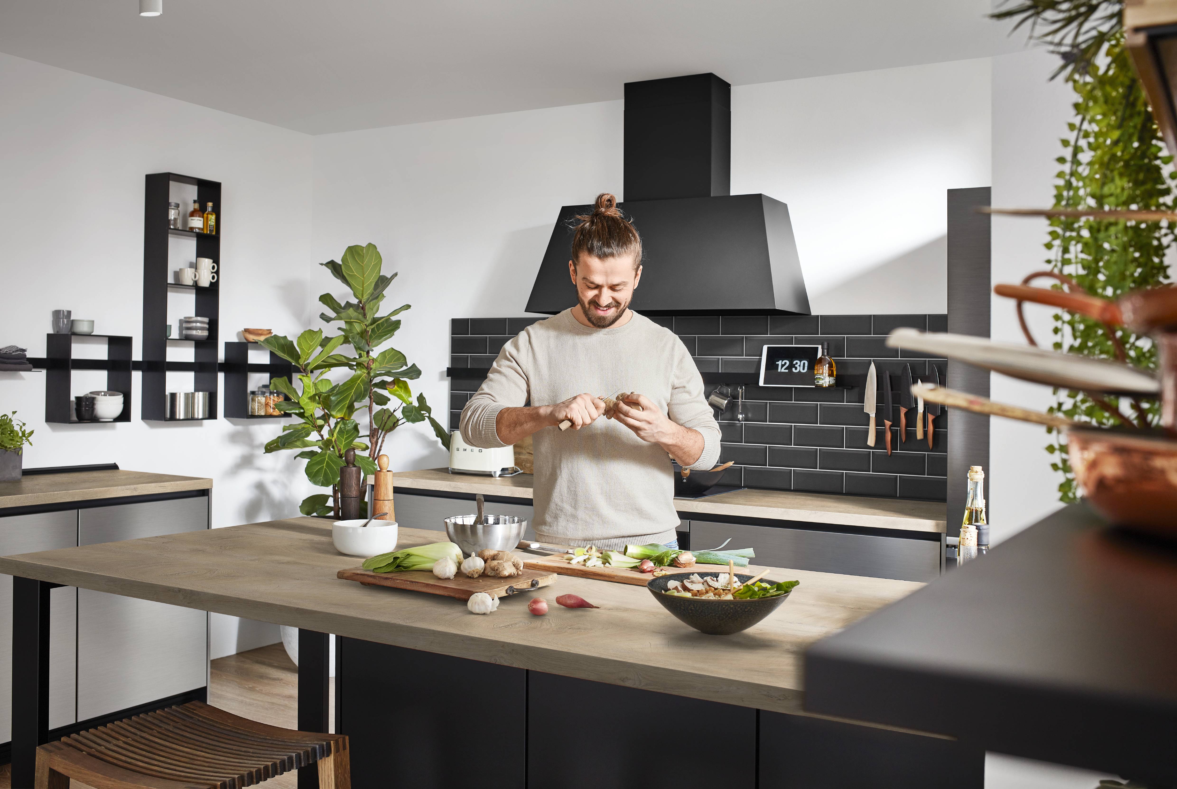 A smiling man with a bun hairstyle preparing vegetables at an oak-topped kitchen island in a matte black and oak kitchen, with black subway tile backsplash, a large black chimney hood, wall-mounted open black shelving, a magnetic knife rail, indoor plants, and a Smeg toaster on the worktop.