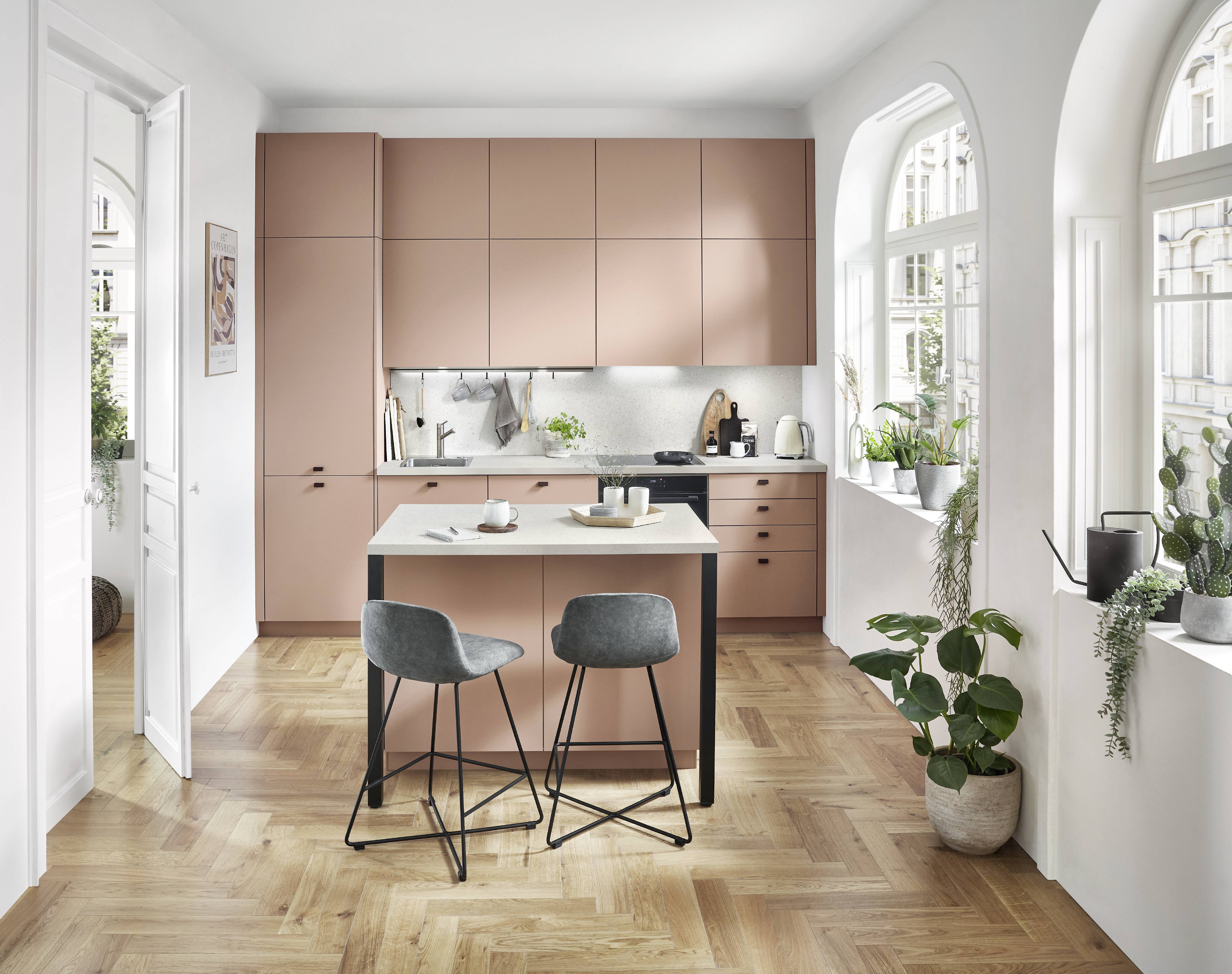 Dusty Rose Matte Kitchen with Herringbone Floor — Bold Colour in a Compact Layout Compact matte dusty rose handleless kitchen with full-height cabinetry, a terrazzo-effect light worktop with a wall-mounted utensil rail, a freestanding white island bar with black metal legs and two grey upholstered bar stools, herringbone oak wood flooring, arched windows, and abundant indoor plants on the windowsill.