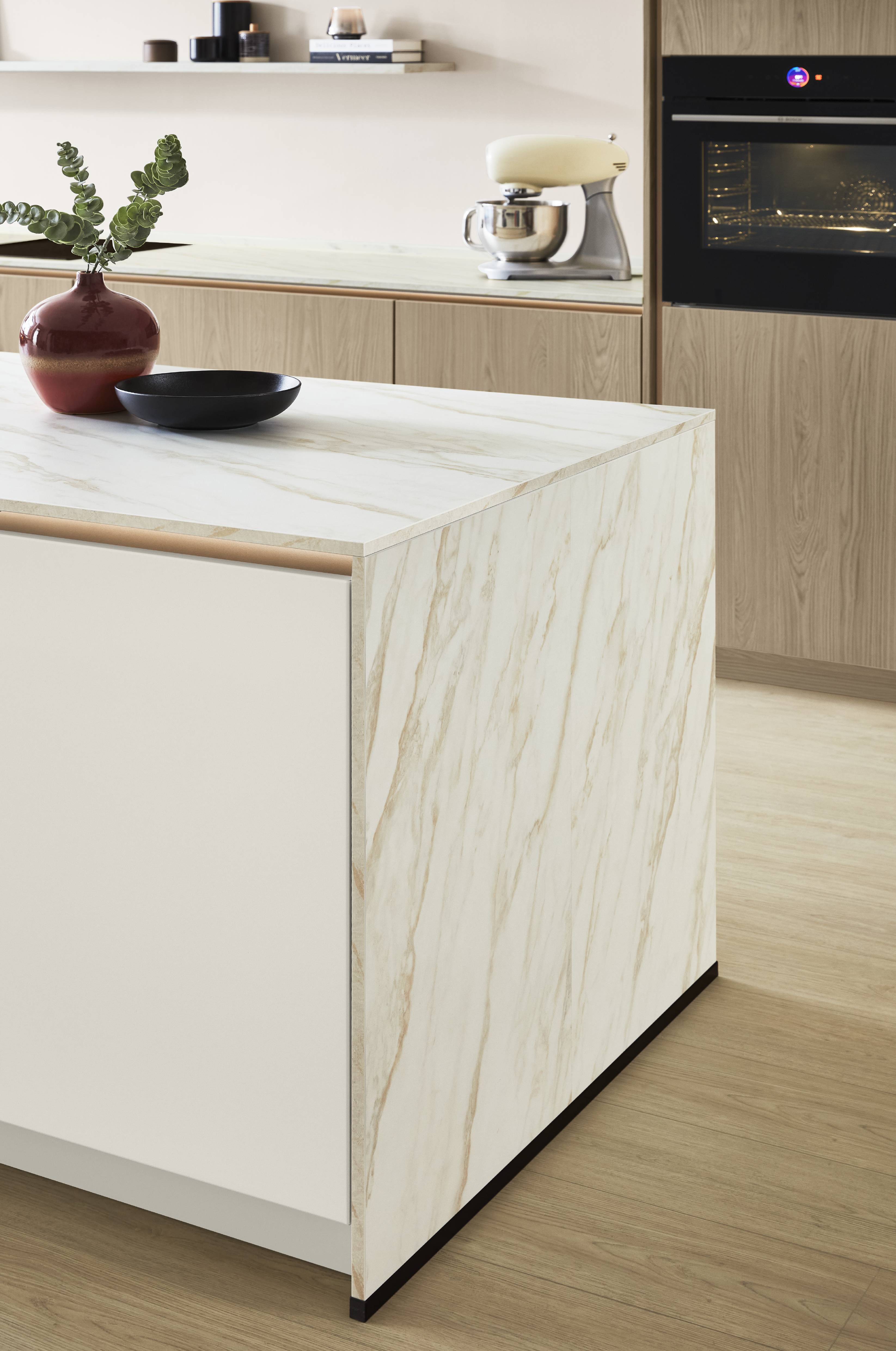 Calacatta Marble-Effect Island End Panel — Full-Height Detail in an Oak and Blush Kitchen Close-up detail of a kitchen island end panel in full-height white Calacatta marble-effect with warm gold veining, wrapping from the matching island worktop to the floor, with a rose gold handle rail, white drawer front, and light oak cabinetry, open shelf, and an active black oven with a cream stand mixer visible in the background.