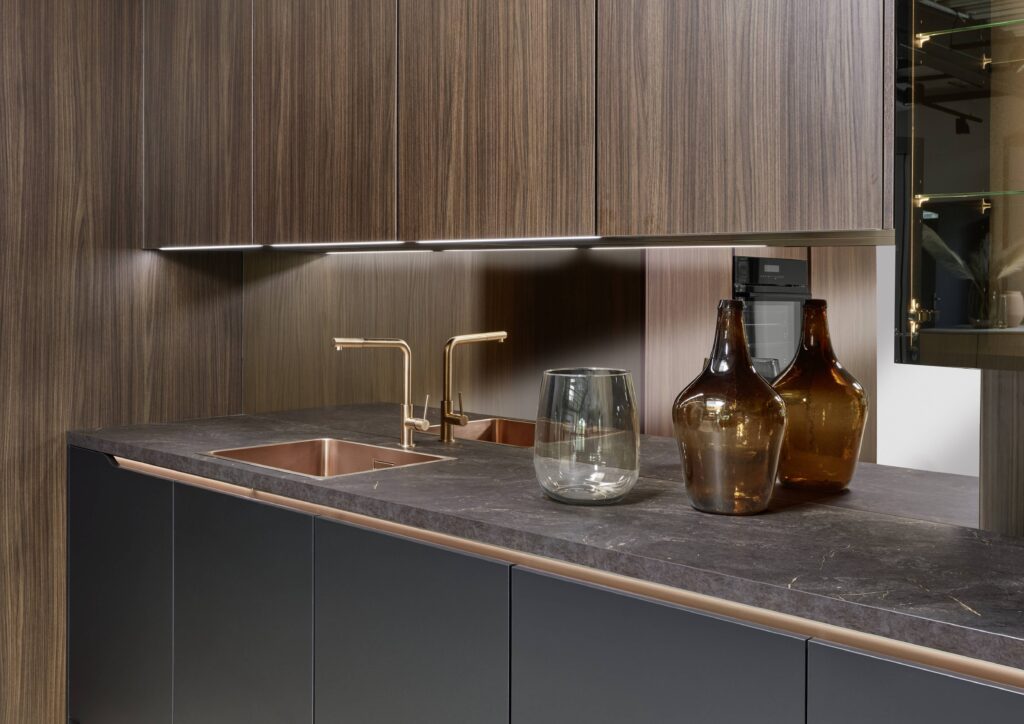 Close-up detail of a luxury kitchen worktop in dark veined marble with a rose gold undermount double sink, brushed brass mixer and hot water taps, matching rose gold cabinet handle strip on graphite lower cabinetry, dark wood-grain upper cabinets, a mirrored backsplash, and amber glass decorative bottles.