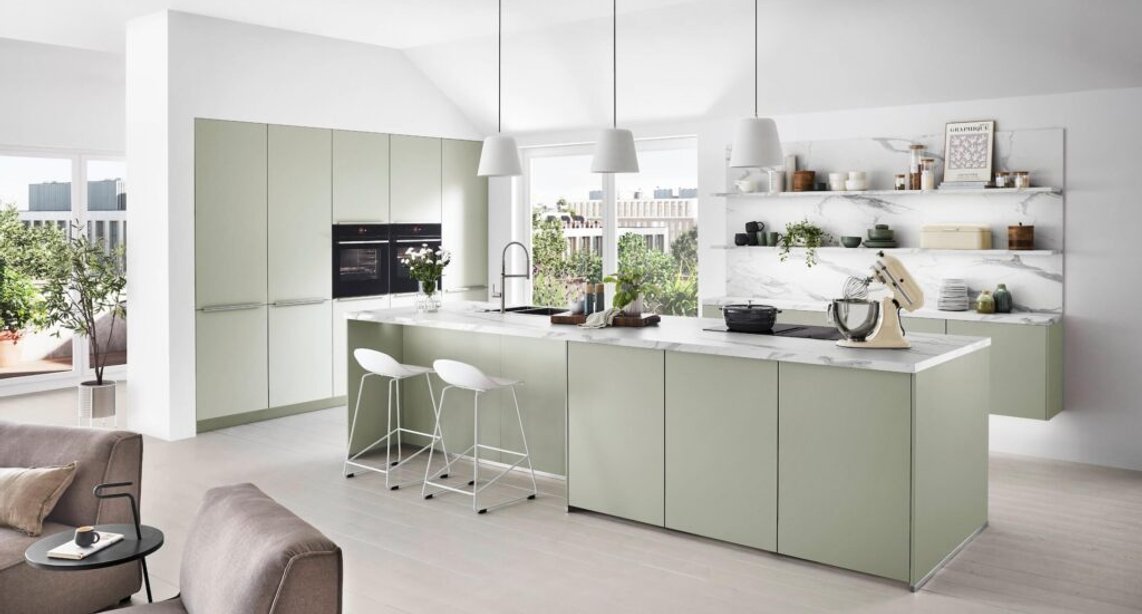 Modular done beautifully — sage green cabinetry, an oversized island, and open marble-backed shelving show what thoughtful kitchen remodeling can achieve.