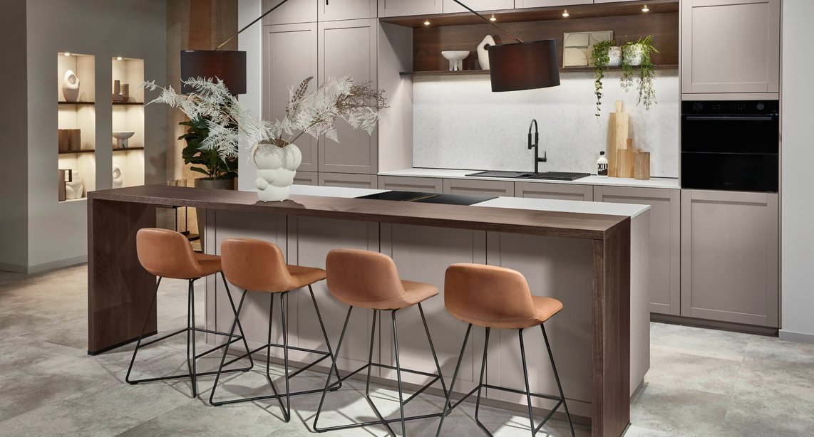 Traditional form, contemporary feeling — a greige shaker kitchen with frame-and-panel cabinetry, a dark walnut island bar, cognac velvet stools, and illuminated display niches proves that classic cabinet styles translate effortlessly into modern living spaces.
