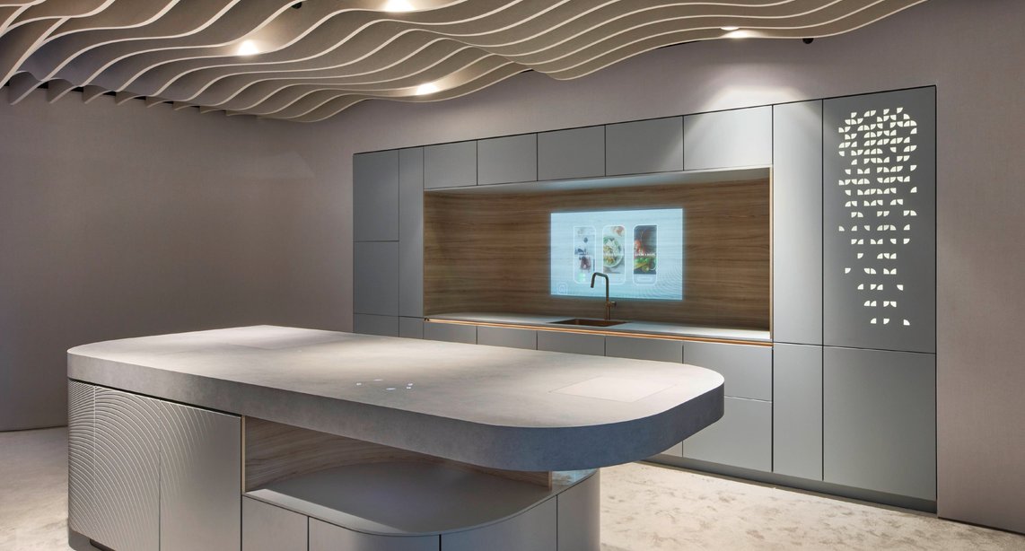 The future of kitchen design, on display — a curved grey island with sculpted relief panels, a smart screen integrated into the backsplash, an Arabic-pattern laser-cut cabinet panel, and a dramatic wave ceiling installation define what boundary-pushing kitchen design looks like today.